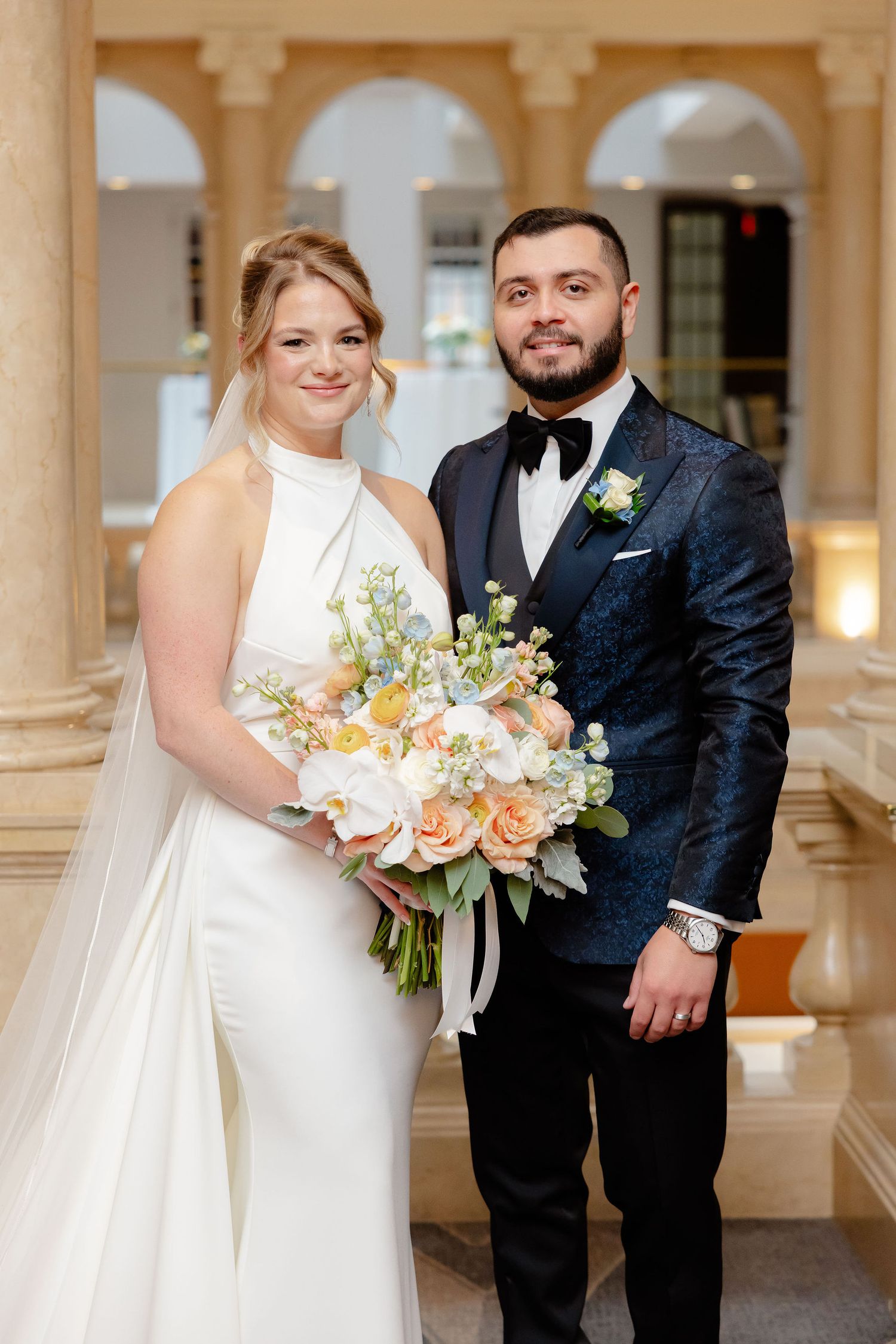 Elegant wedding couple poses in formal attire with peach and white floral bouquet in a columned hallway.