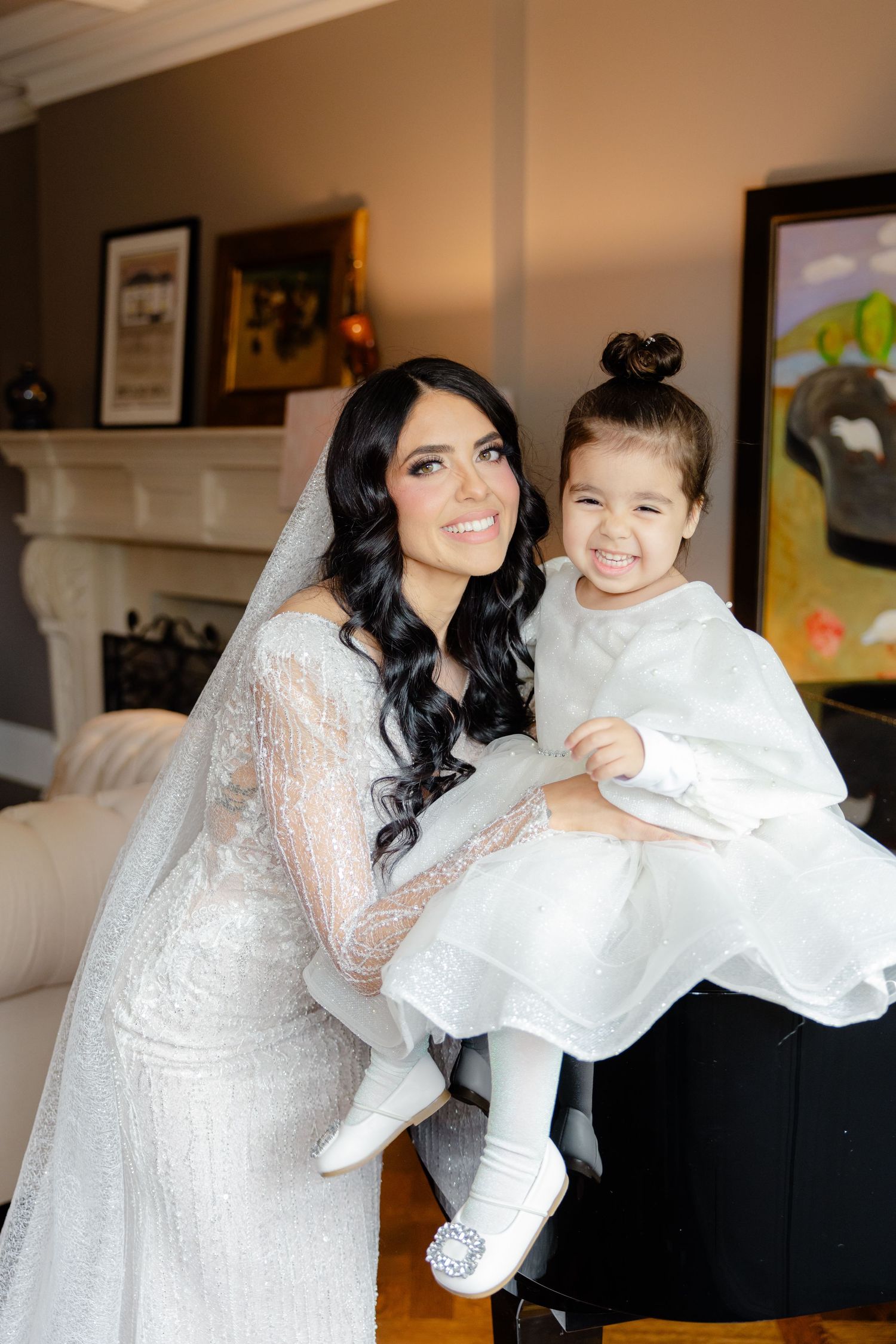 A bride in white lace and young attendant in matching white dress pose by a grand piano.