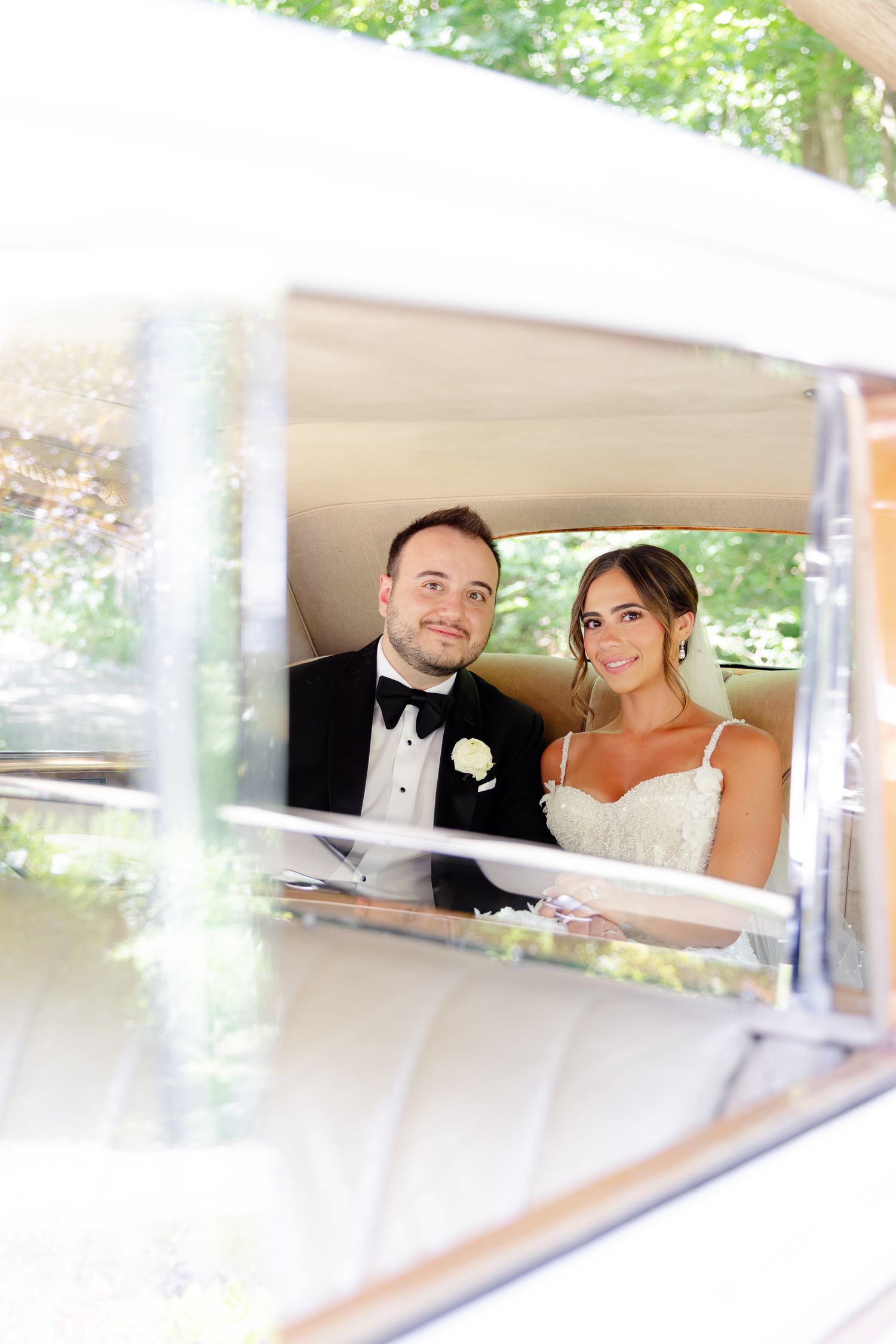 A bride and groom sit together in a vintage car on their wedding day, viewed through the car's window.
