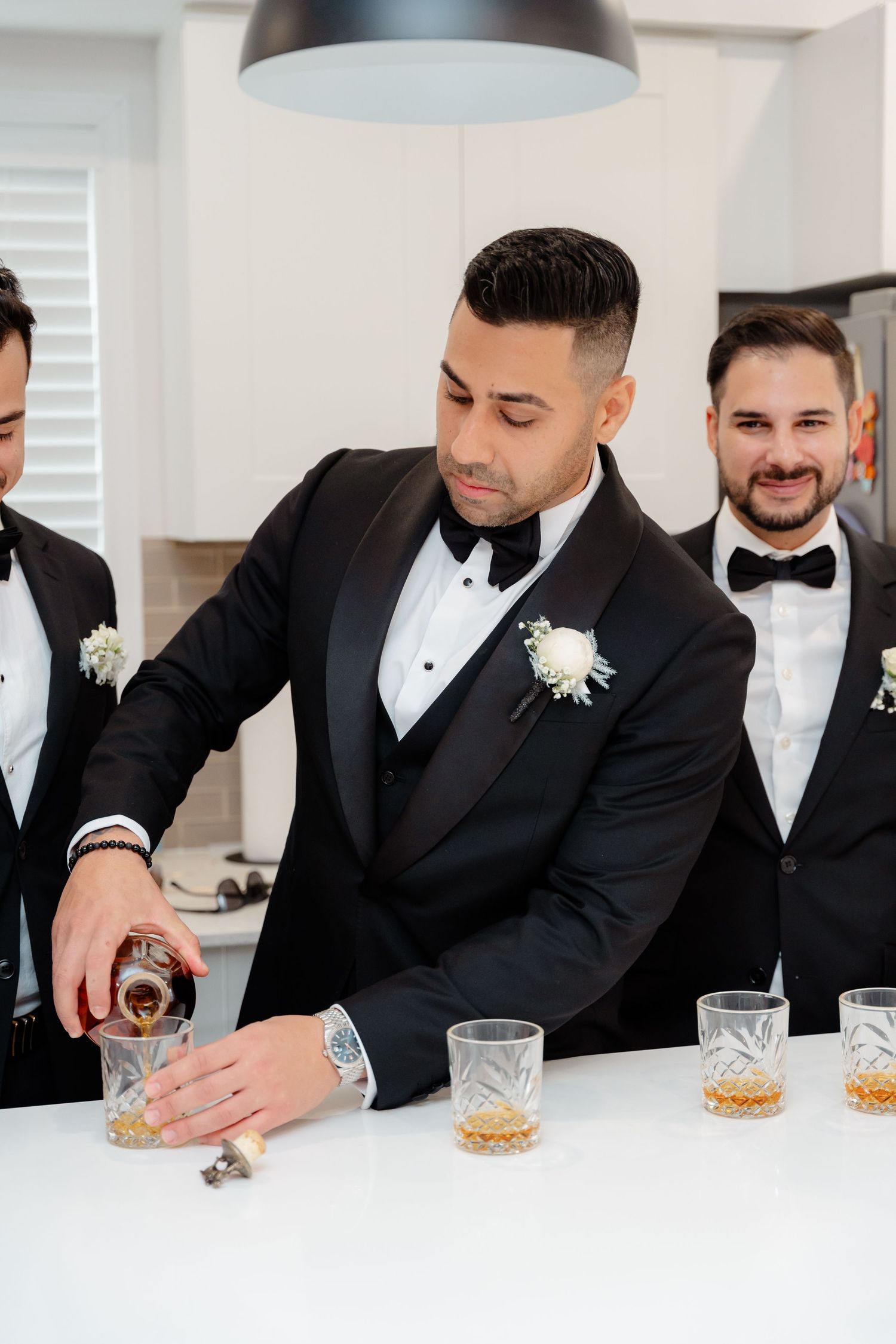 Groomsmen pour drinks at modern white kitchen counter under pendant lights.