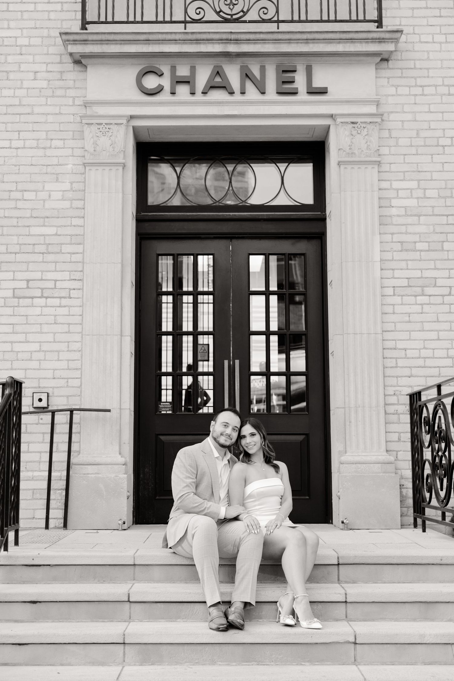 Black and white photograph of a couple seated on the steps of a Chanel boutique entrance.