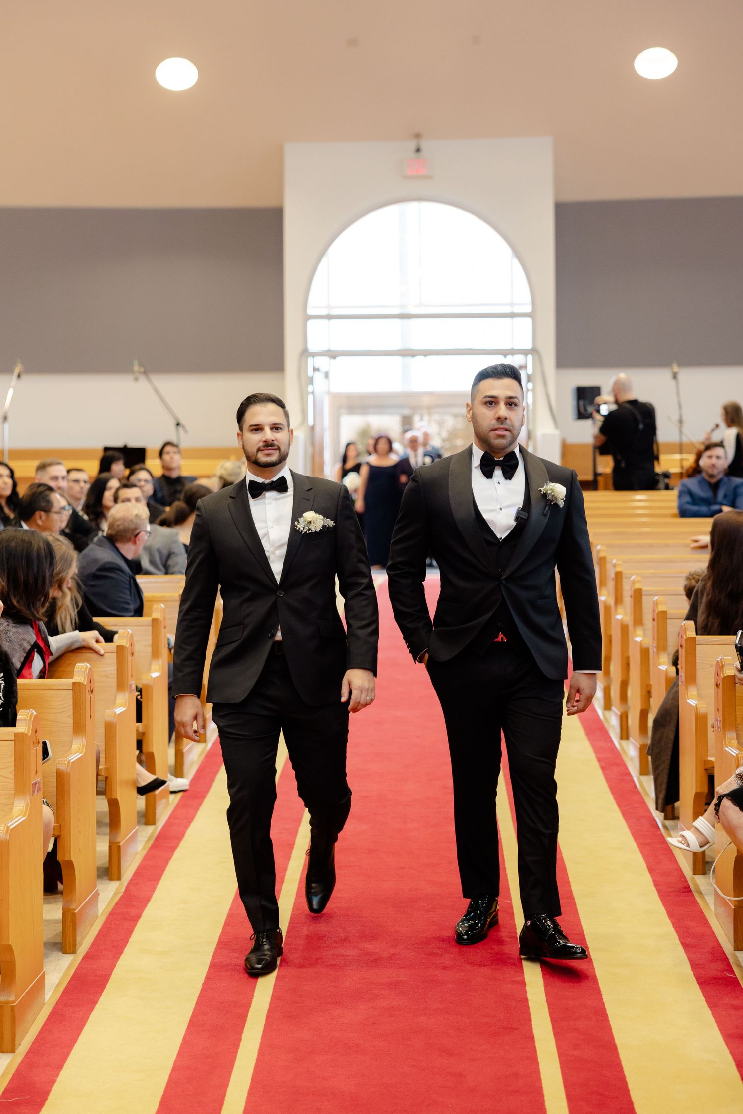 Two people in black tuxedos walking down a red carpeted church aisle during a ceremony.
