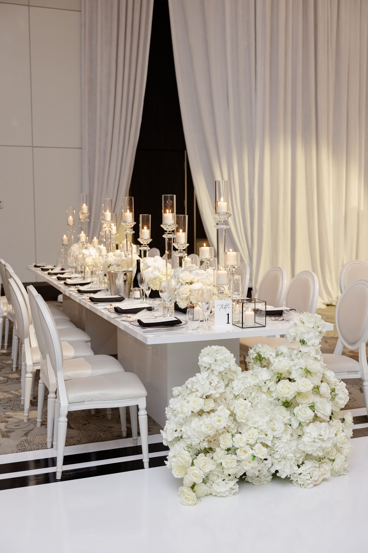 Elegant wedding reception table with white hydrangeas, crystal candle holders and white chairs against dark backdrop.