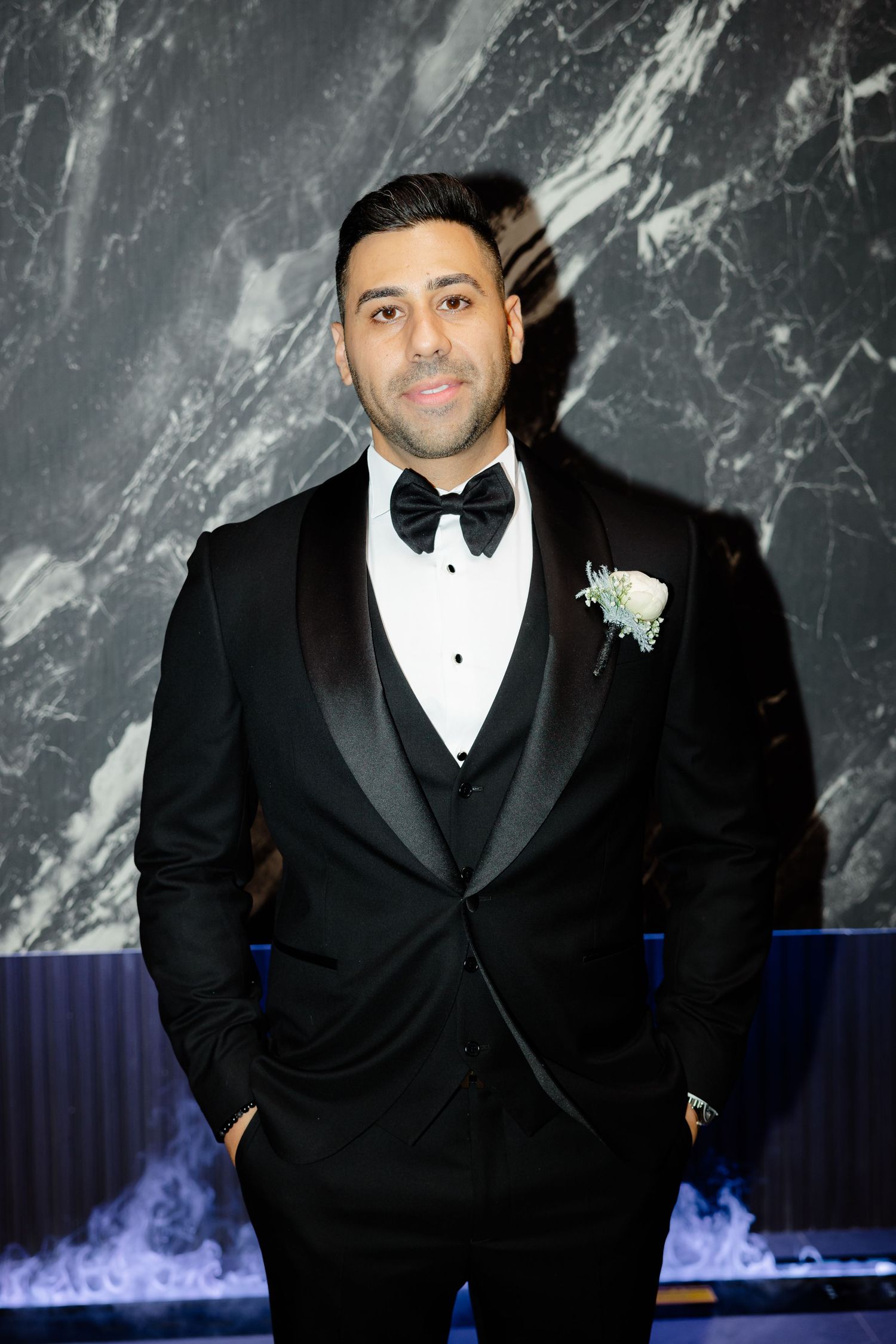 Groom in classic black tuxedo poses against marble wall with blue lighting accent.