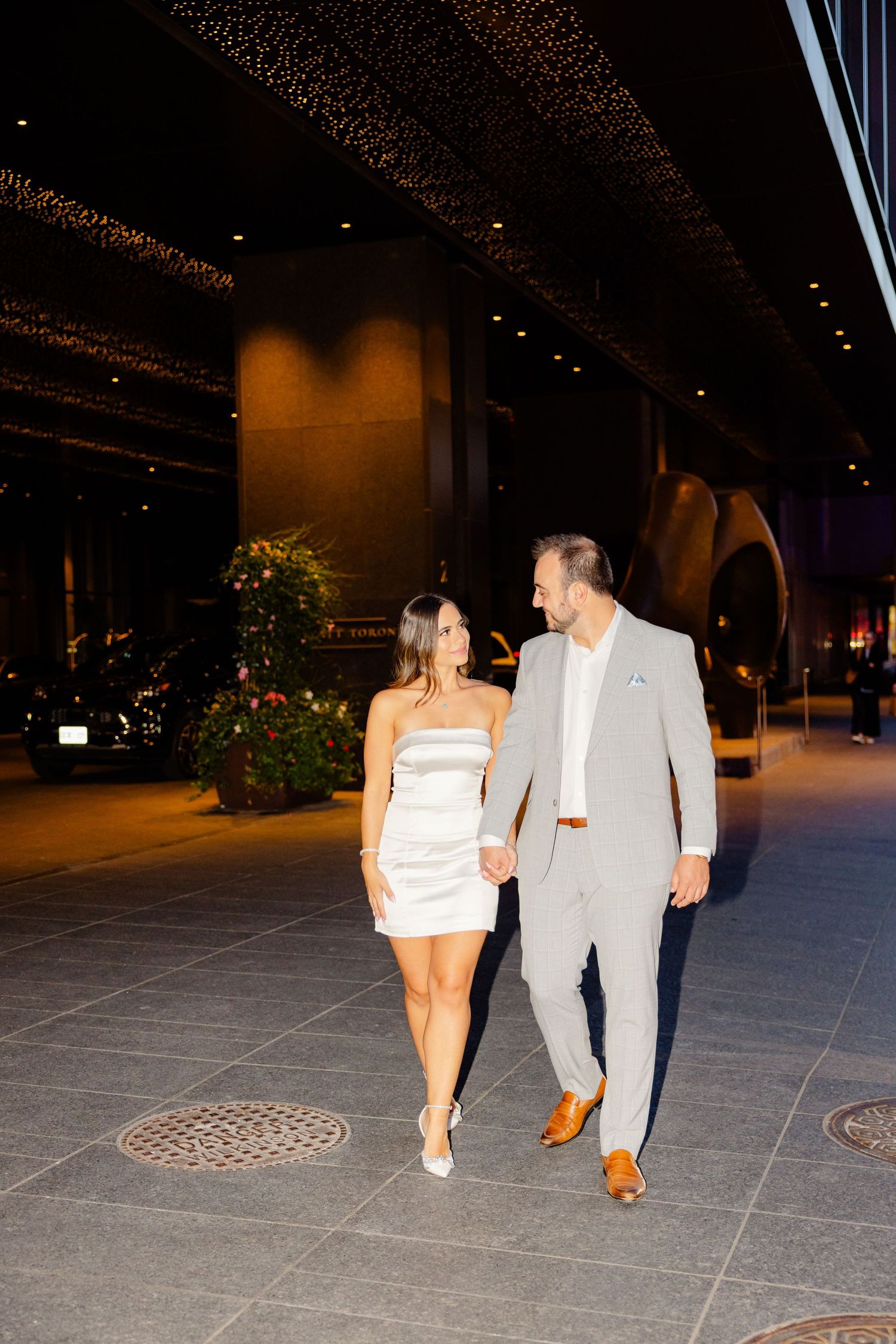 Well-dressed couple walking hand in hand on outdoor plaza at night.