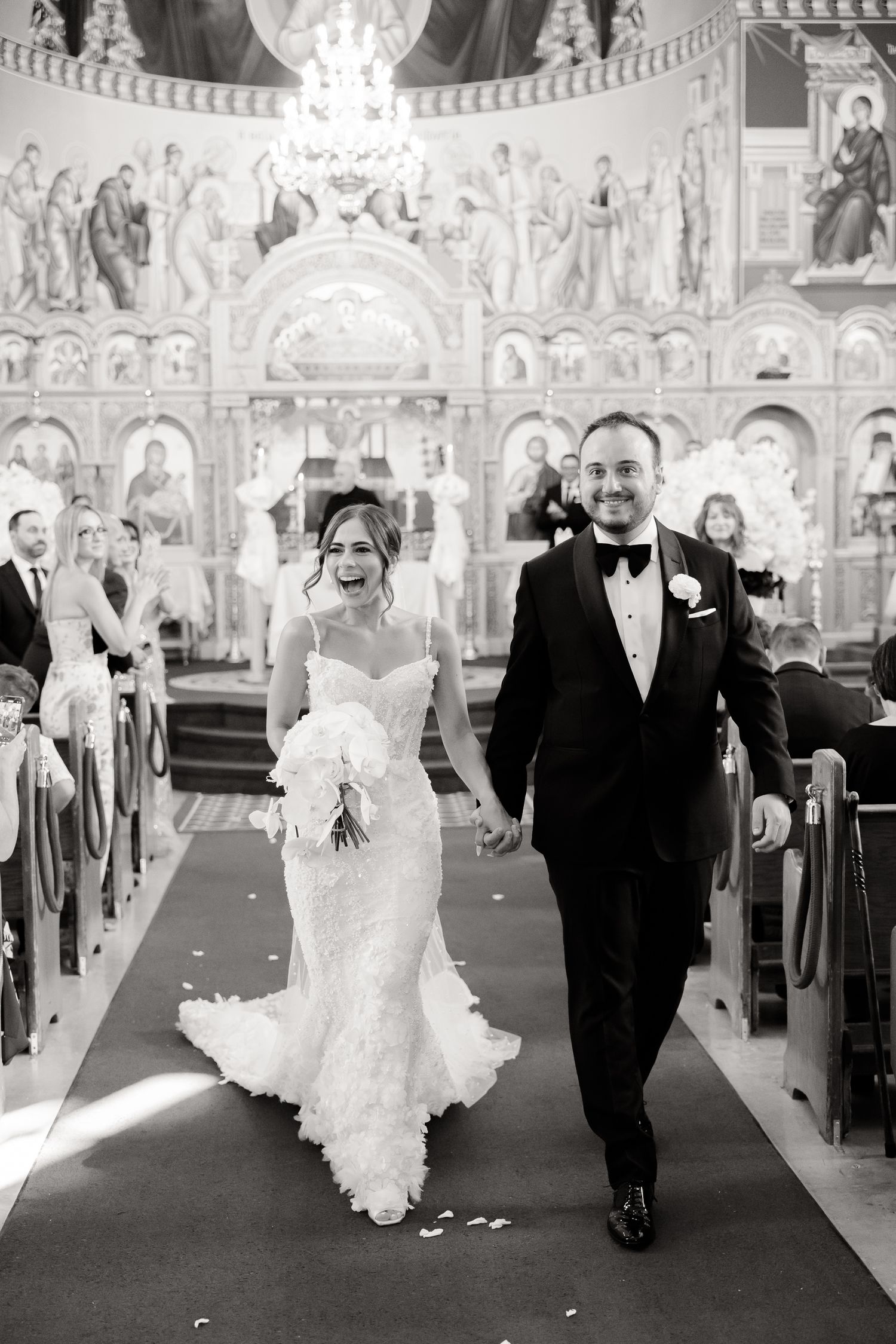 A couple walks down the aisle after their wedding ceremony in an ornate Orthodox church with beautiful frescoed walls.