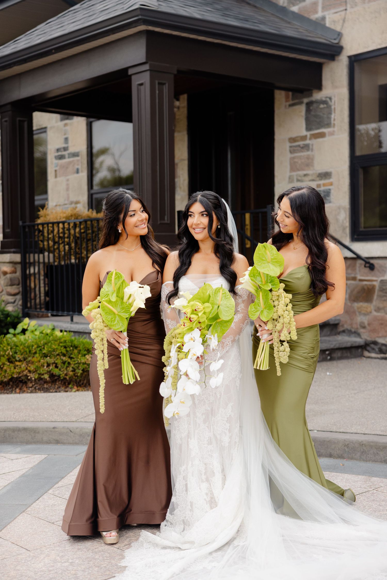 A bridal party wearing brown and olive green dresses stands together while holding bouquets outside a stone building.