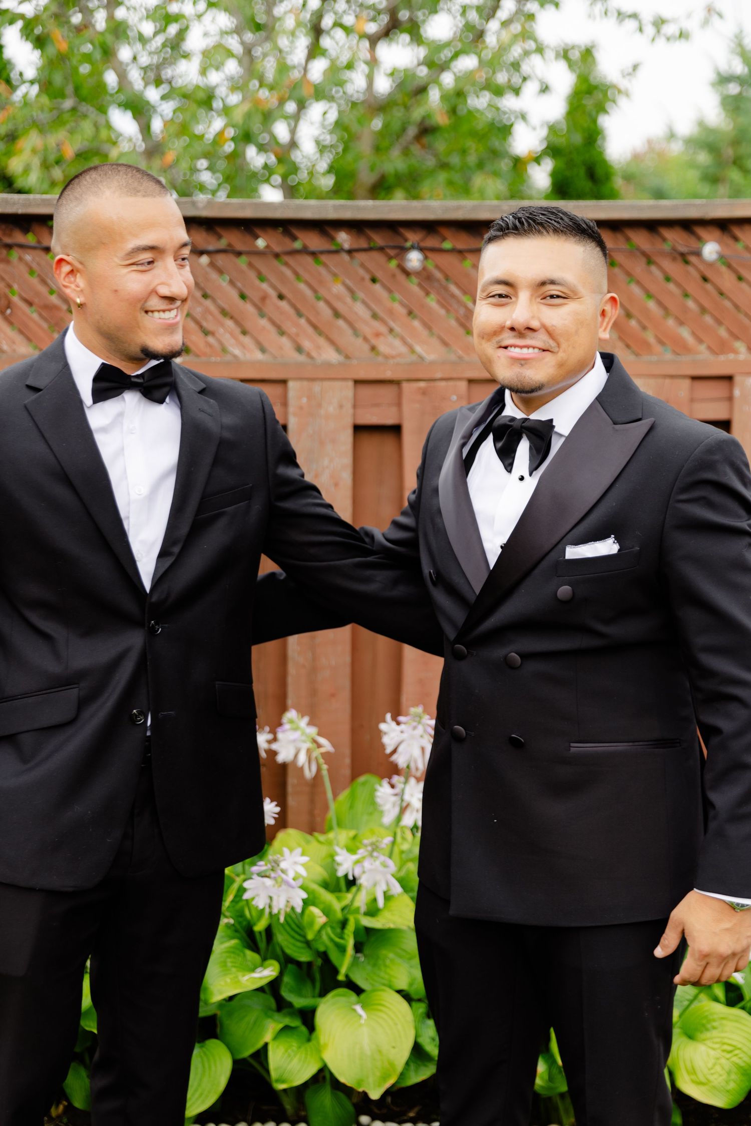 Two men in black tuxedos share a joyful moment together in front of a garden fence.
