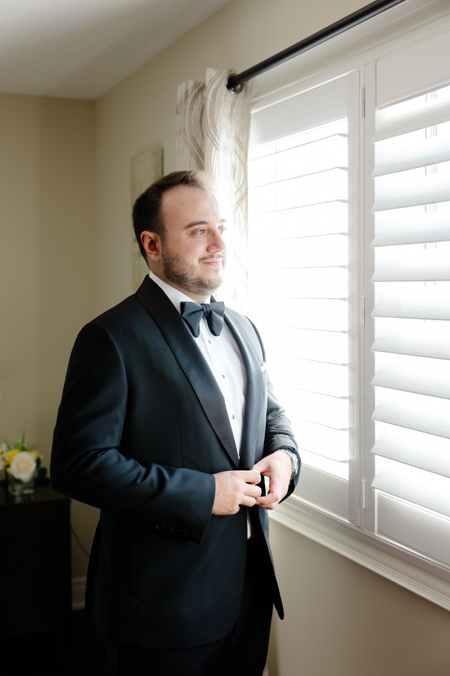 A person in a black tuxedo stands by a window with white shutters looking out into natural light.