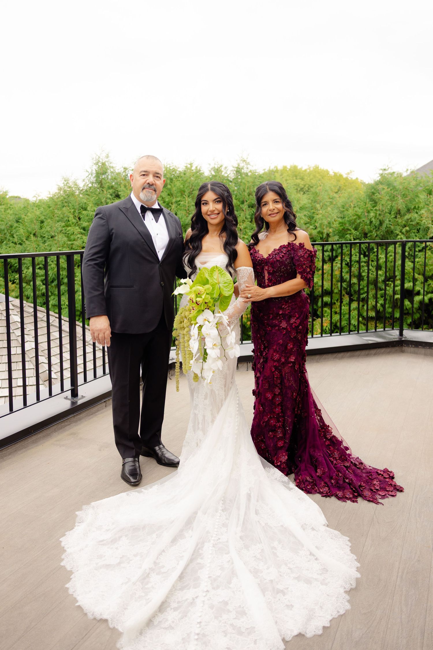 Wedding party poses on outdoor terrace with elegant burgundy and white gowns against wrought iron railing.