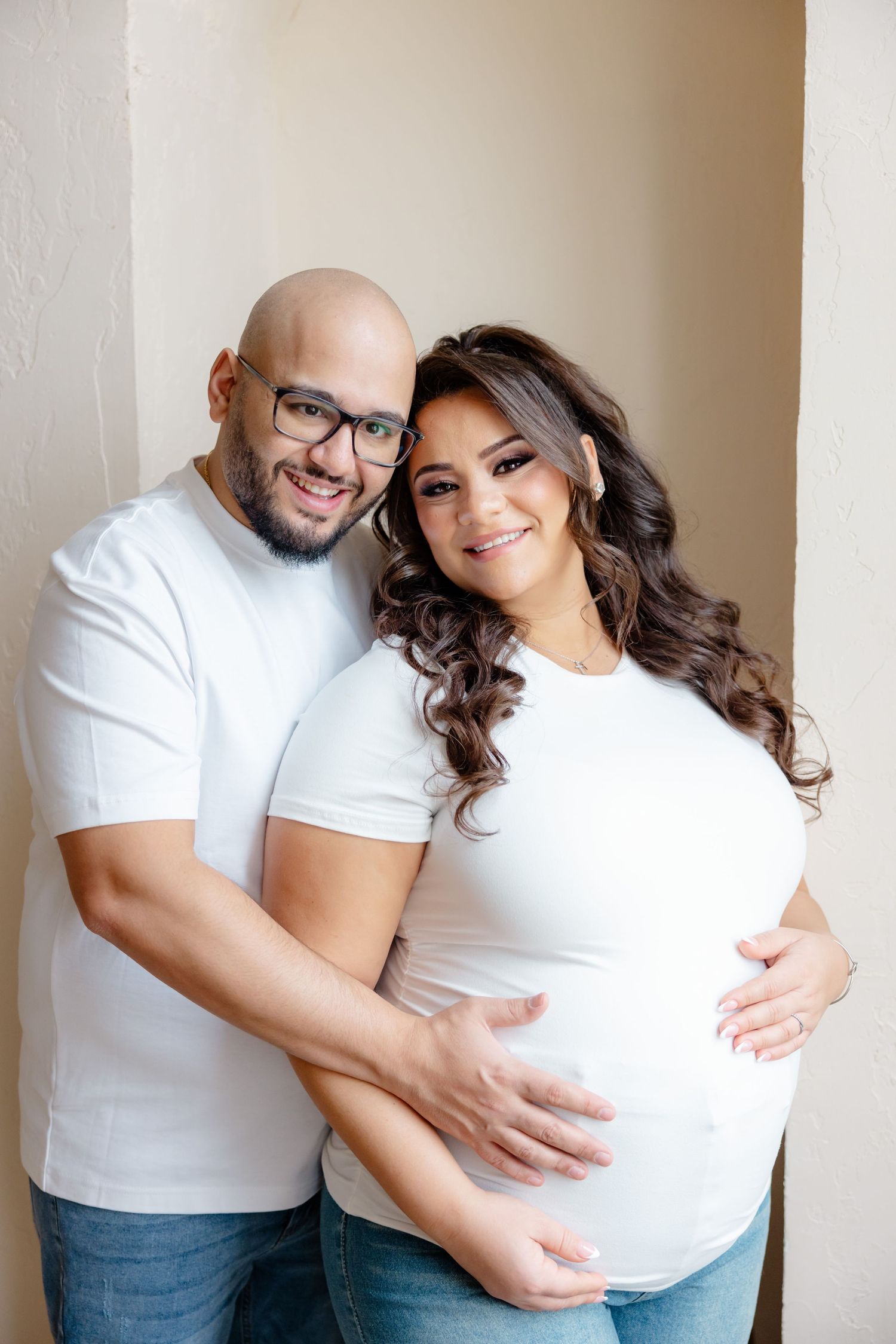 Expectant couple wearing matching white tops embrace while standing against beige wall during maternity photoshoot.