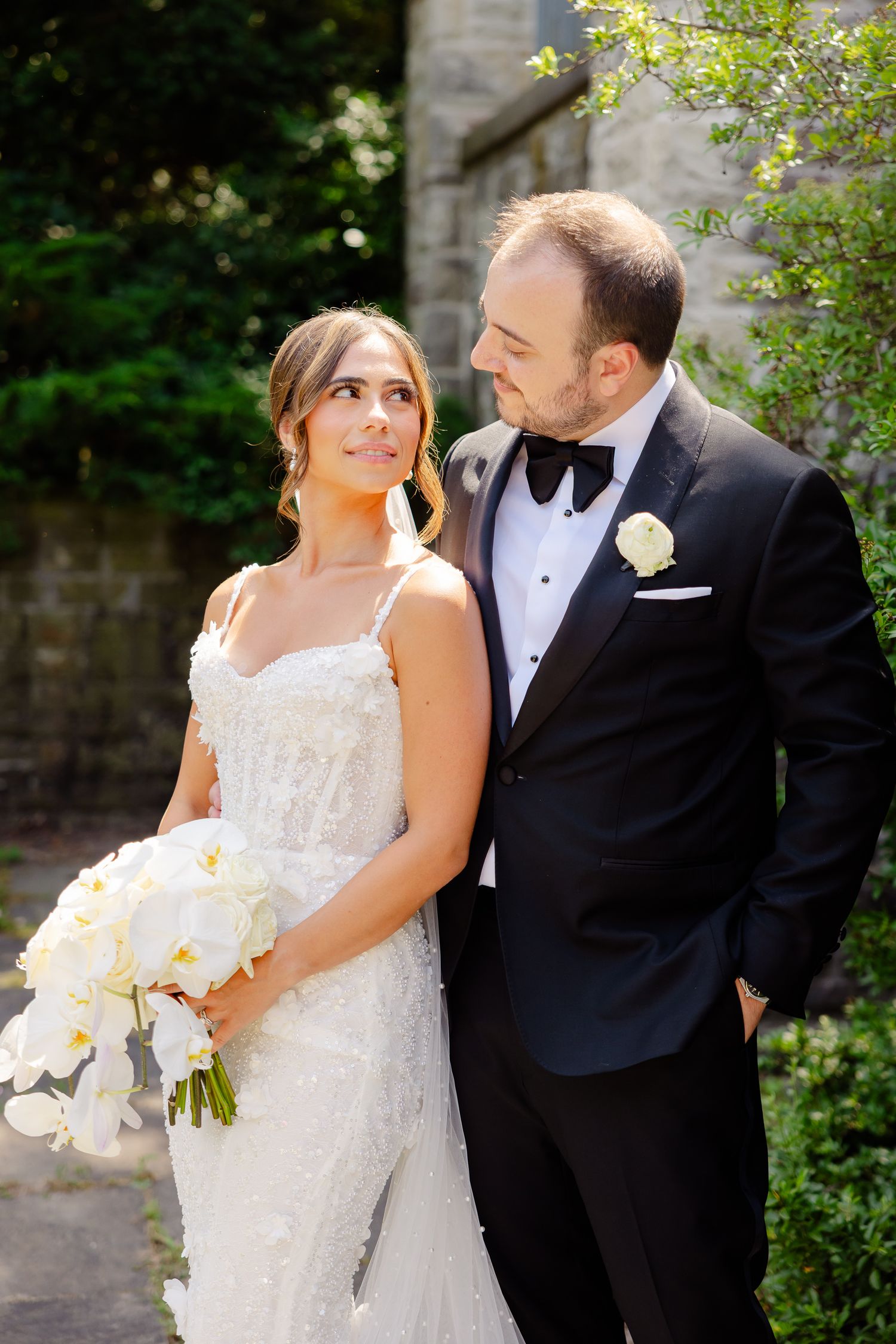 A bride in a lace wedding gown holds a white orchid bouquet while sharing a moment with her groom in a black tuxedo.