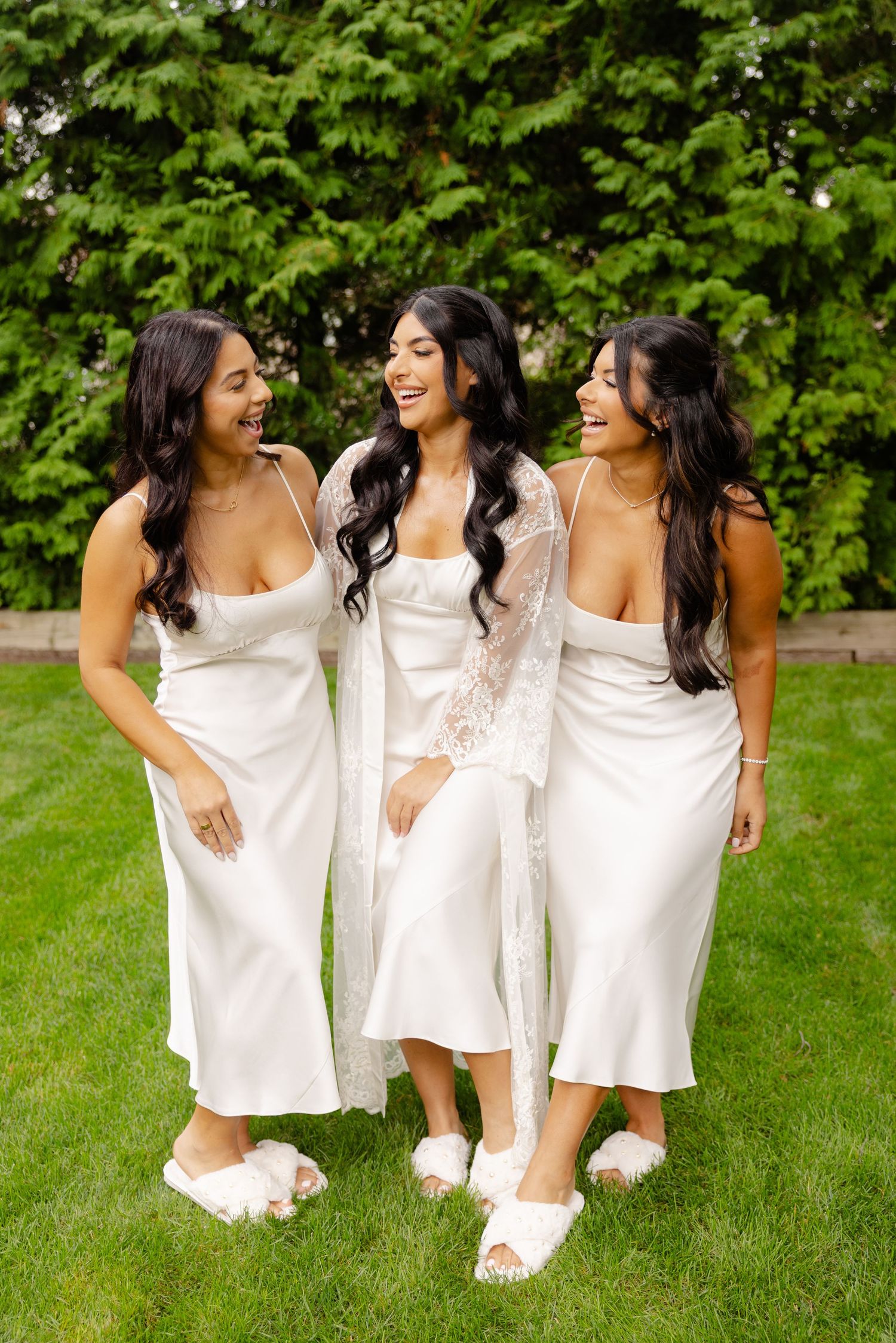 Three women in matching white dresses strike playful poses on grass with green bushes behind them.