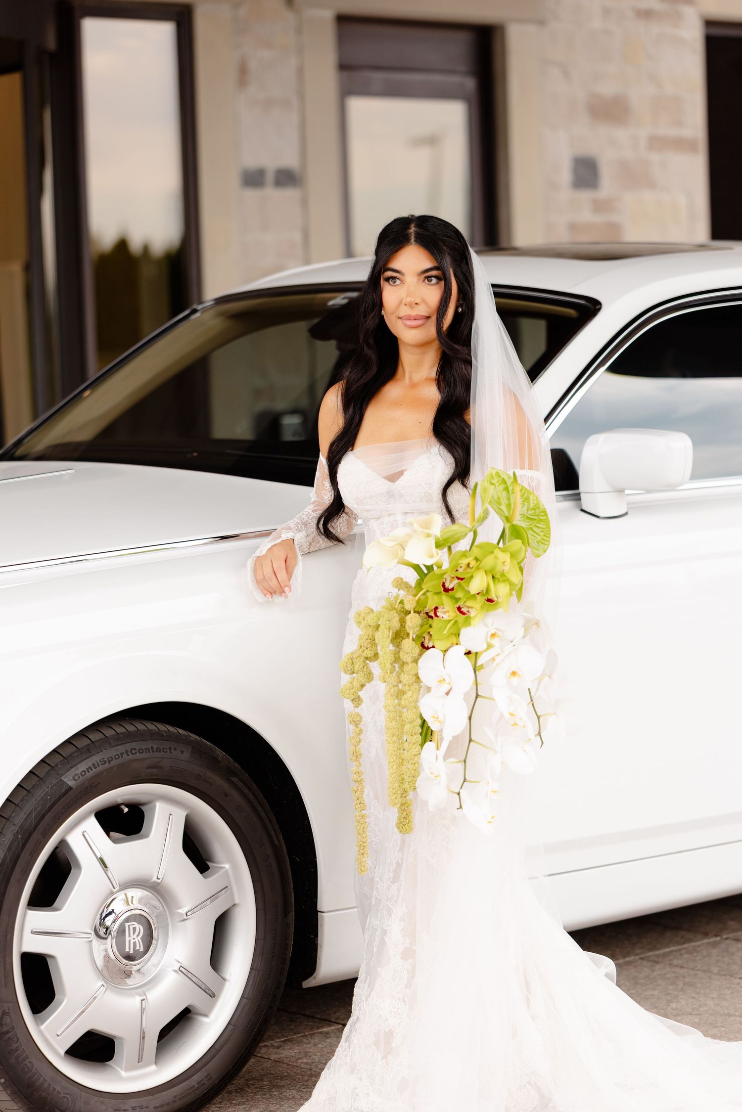 Woman in white dress holding green flowers poses beside luxury white car with Mercedes alloy wheels.