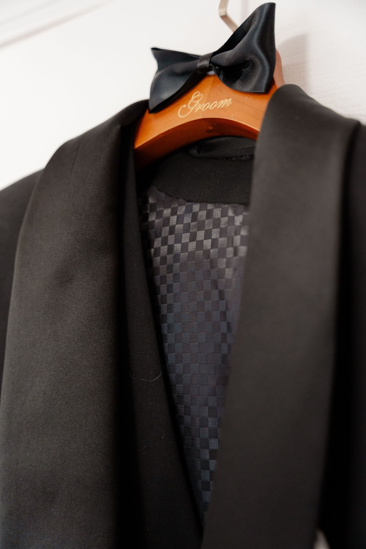 Detail shot of black tuxedo jacket with checkered lining and bow tie on wooden hanger.