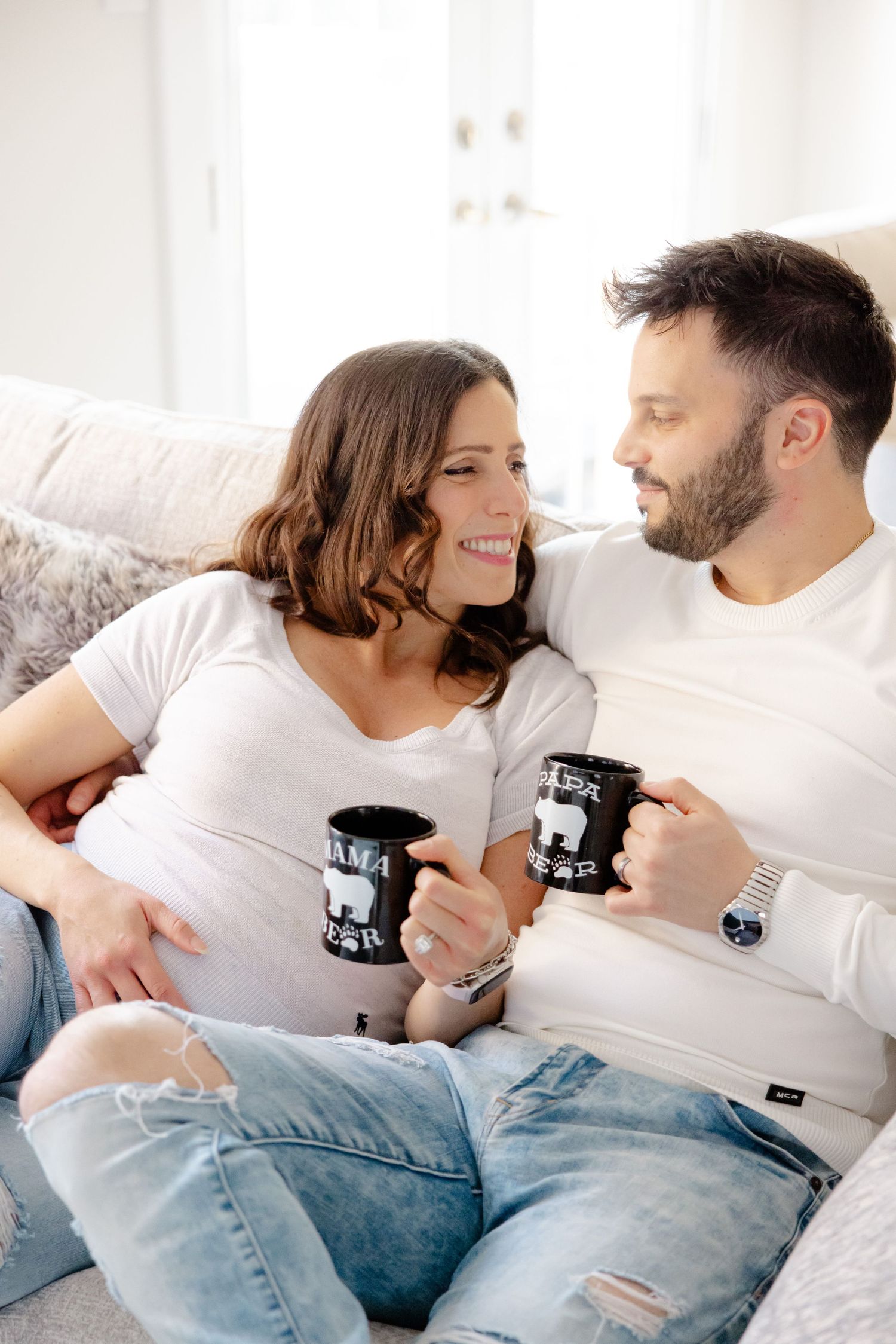 A couple relaxes on a couch, smiling at each other while holding coffee mugs in a bright, cozy room.