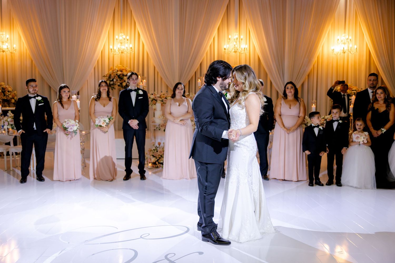Couple shares their first dance surrounded by wedding party in elegant ballroom setting.