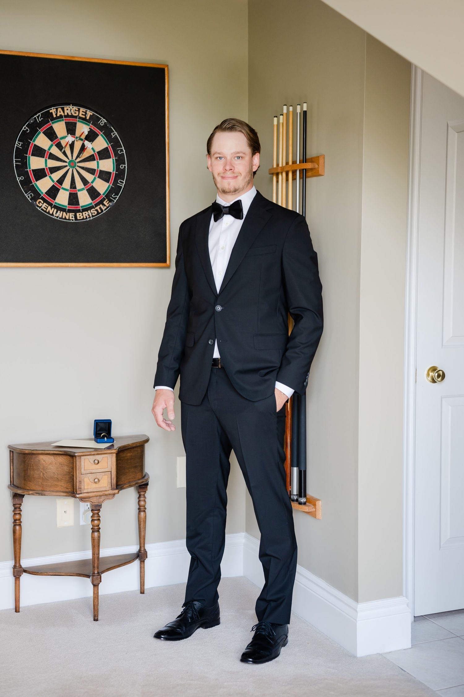 Person in black tuxedo standing by dartboard and antique table in formal setting.