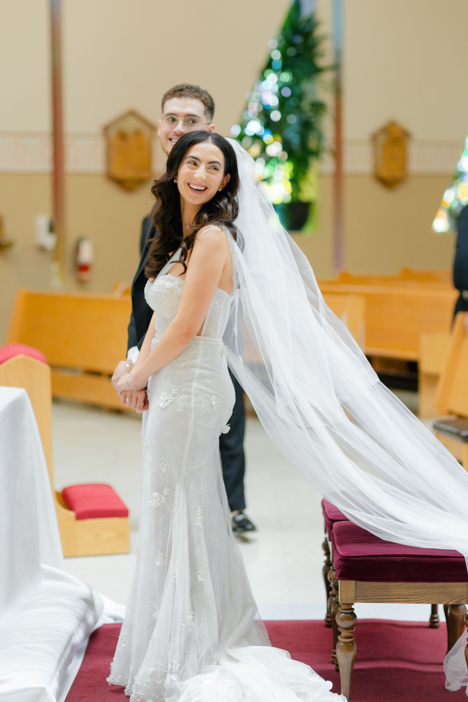 A bride in a fitted lace wedding dress with a flowing veil poses in a church.