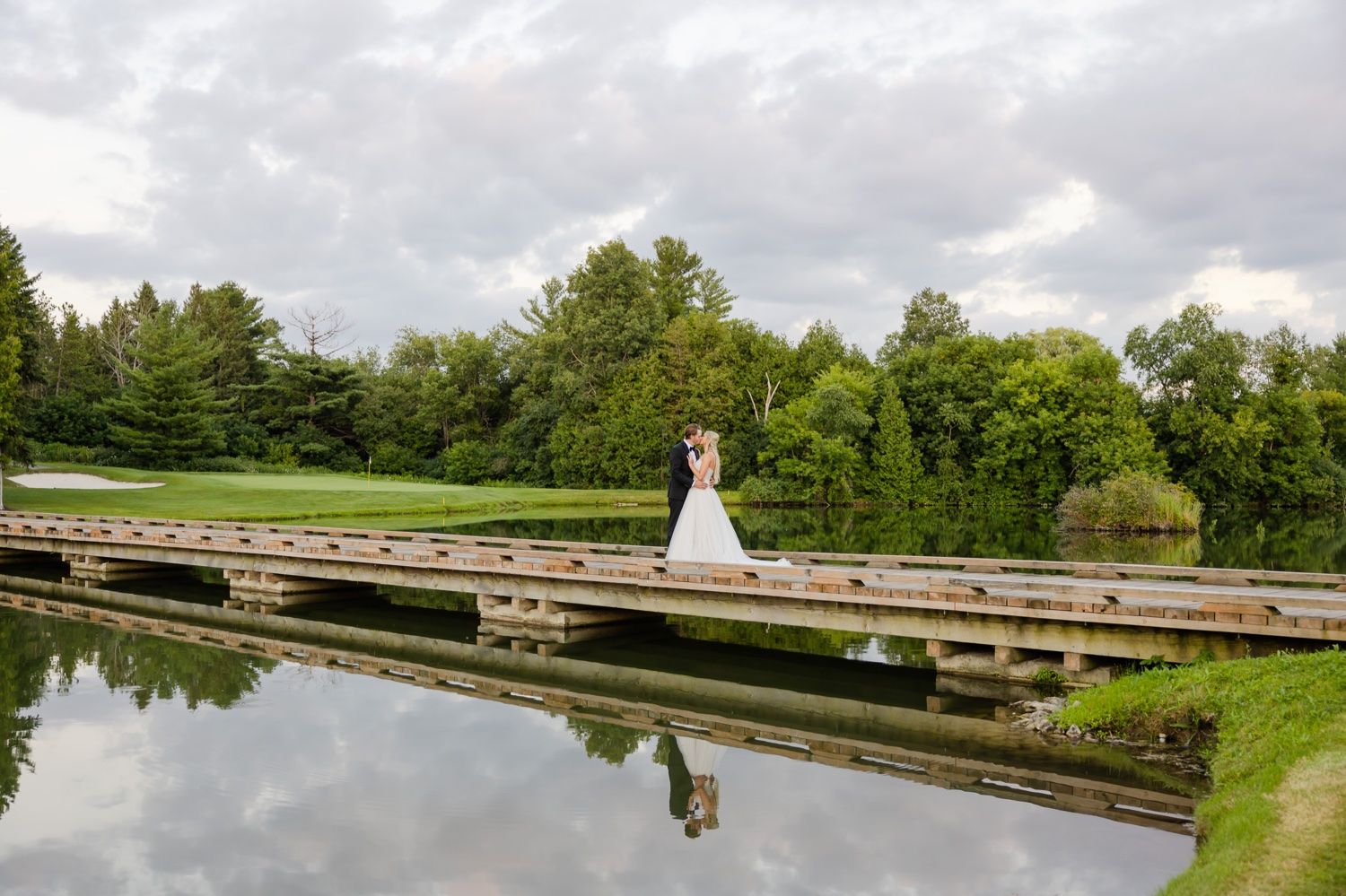 Beautiful wedding dress silhouetted against green trees and reflected in still water by wooden dock.