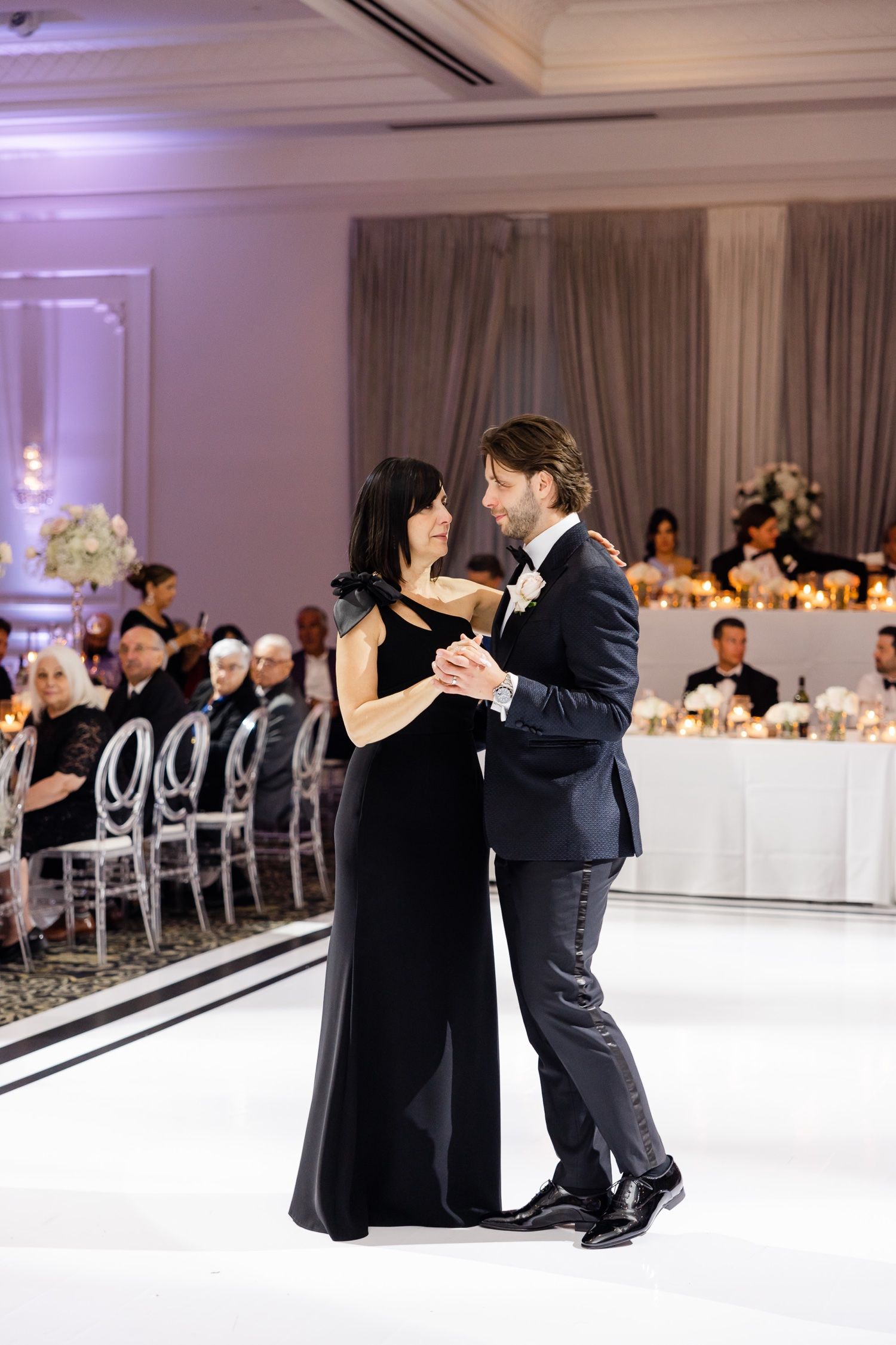 Elegant couple shares intimate dance on white dance floor with reception guests seated nearby.