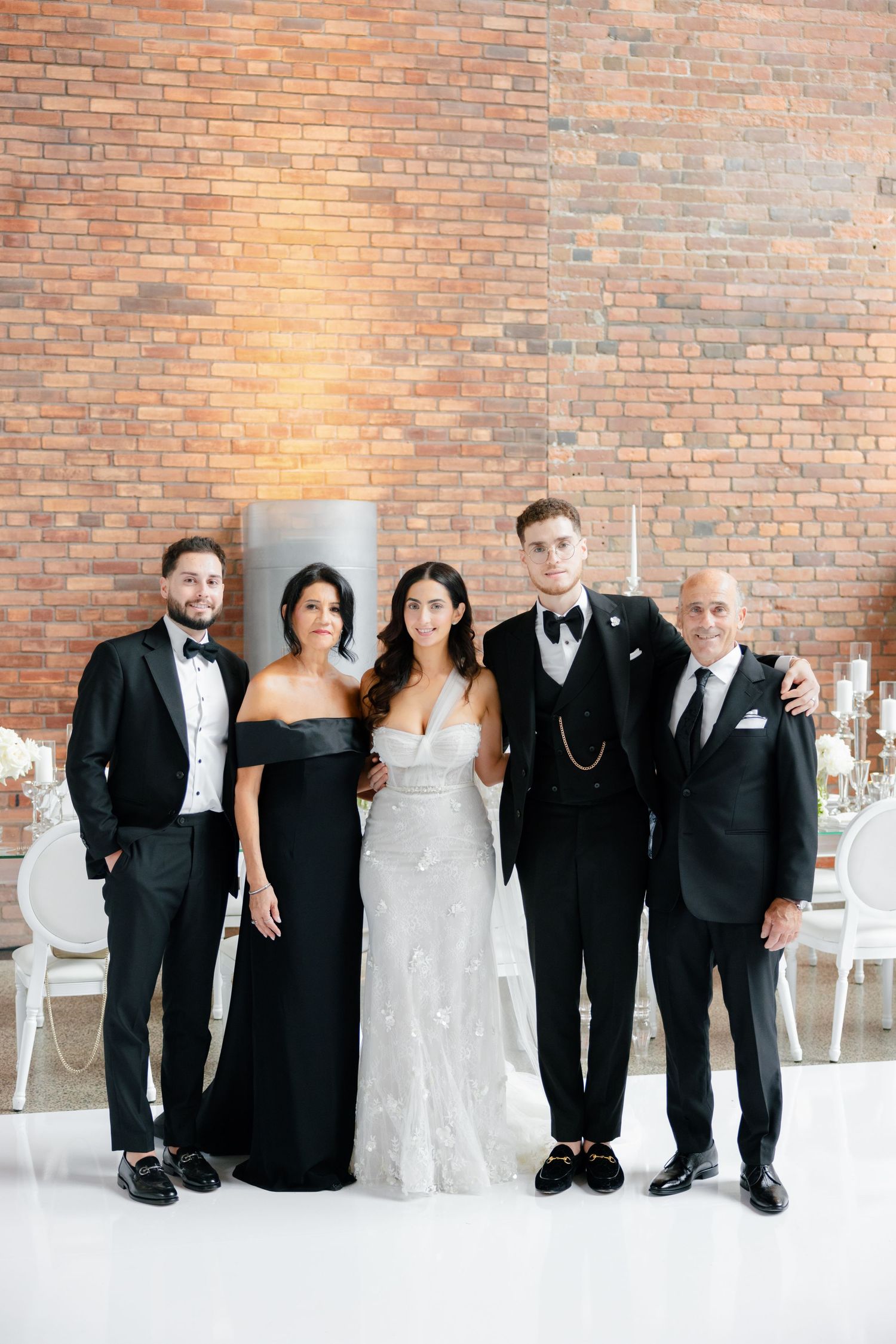 Wedding party in formal black and white attire lined up against rustic brick wall backdrop.