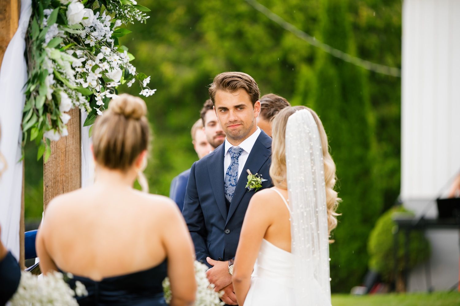 An outdoor wedding ceremony takes place under a wooden arch adorned with white flowers and greenery.