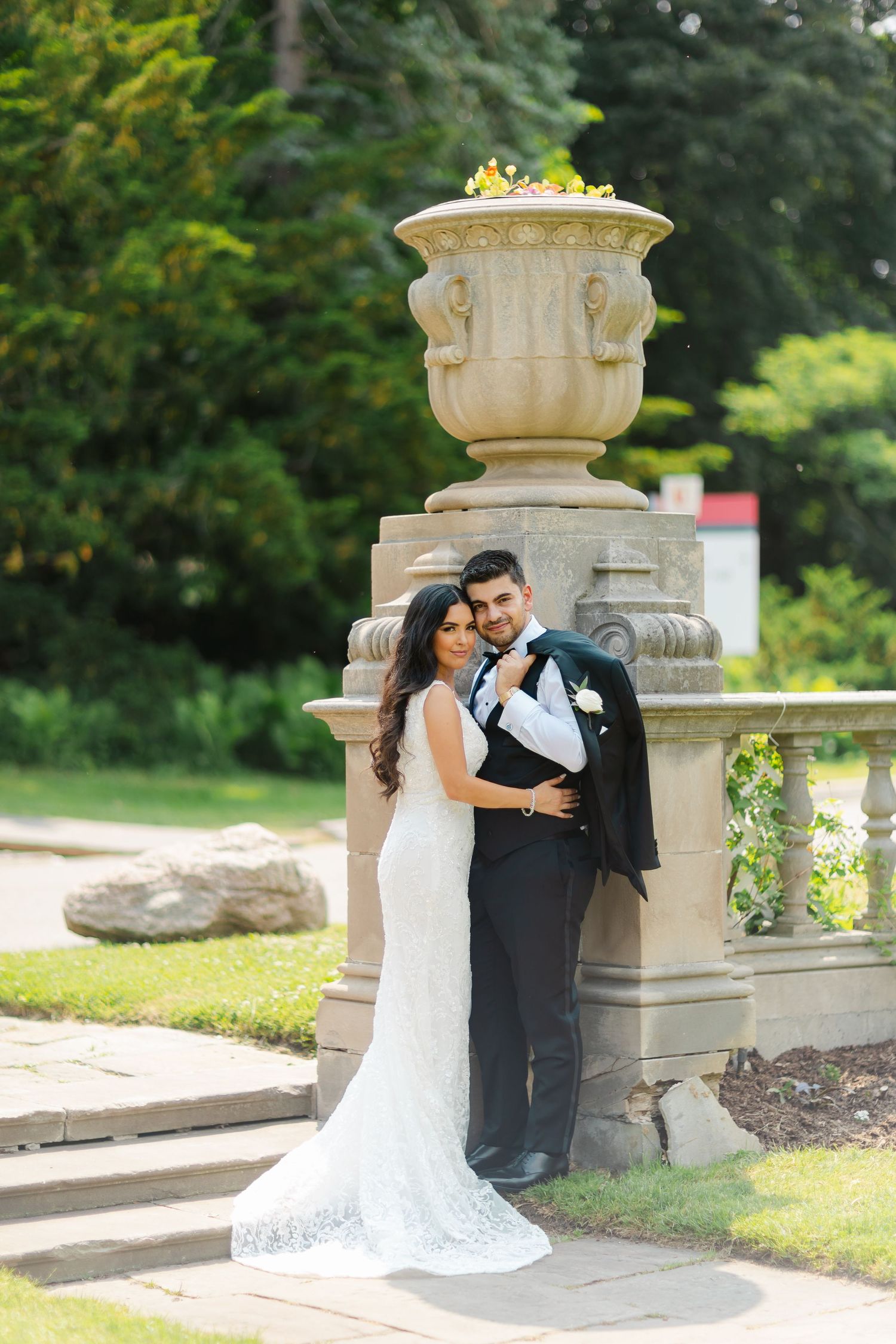 Elegant wedding couple poses by stone architectural feature in lush garden setting.