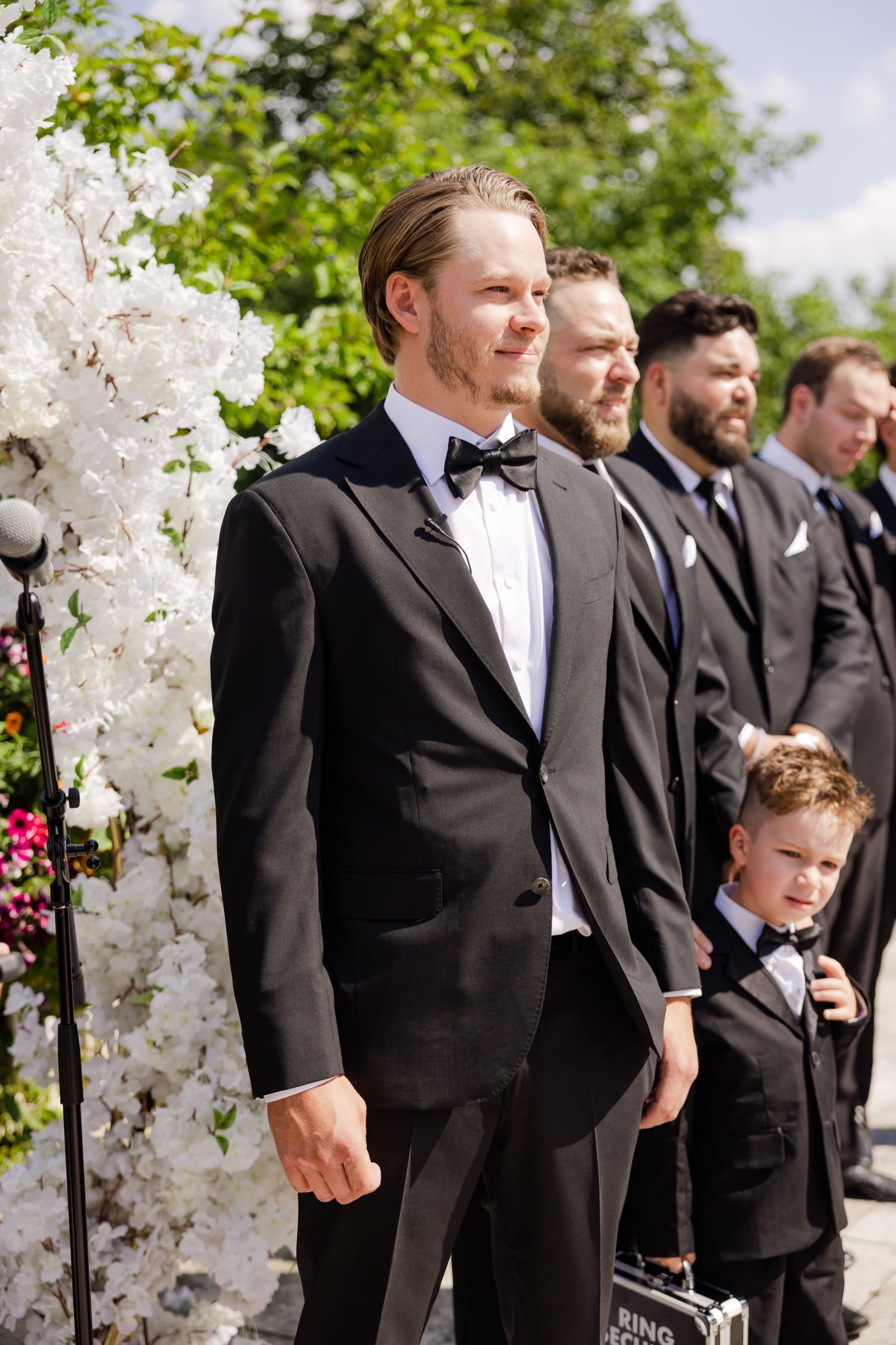 Groomsmen in black tuxedos standing at floral-adorned wedding ceremony altar.