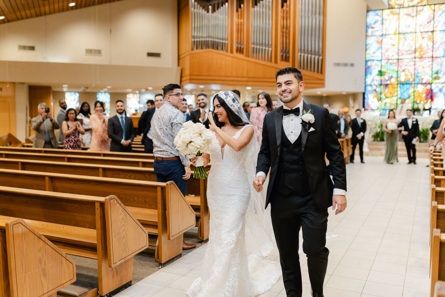 A wedding couple walks down the church aisle with wooden pews and stained glass windows in the background.