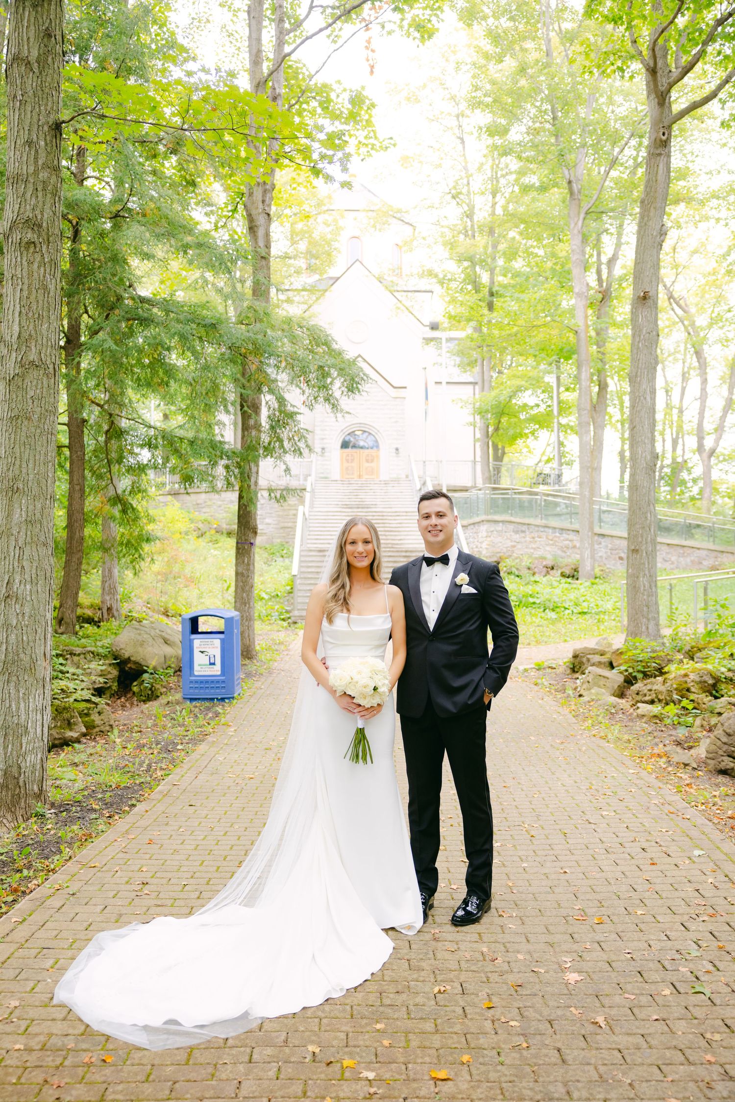 Newlyweds pose on a brick pathway with a white chapel visible through the trees.