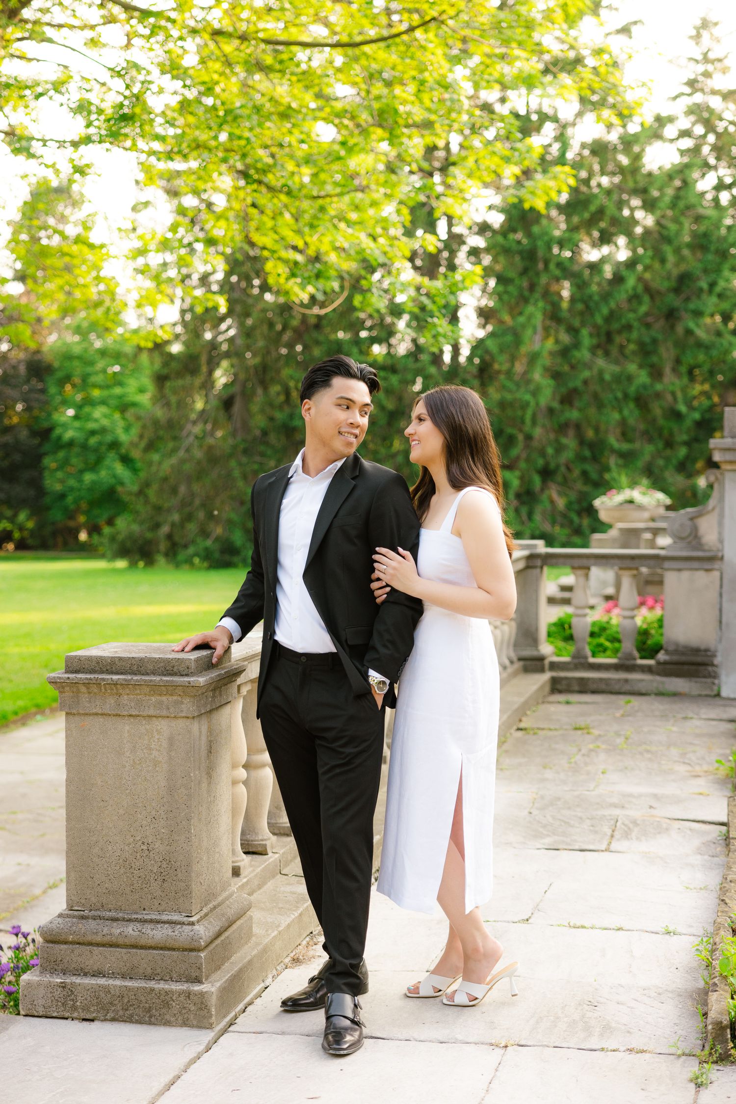 Couple in formal attire sharing tender moment by stone railing in lush garden setting.