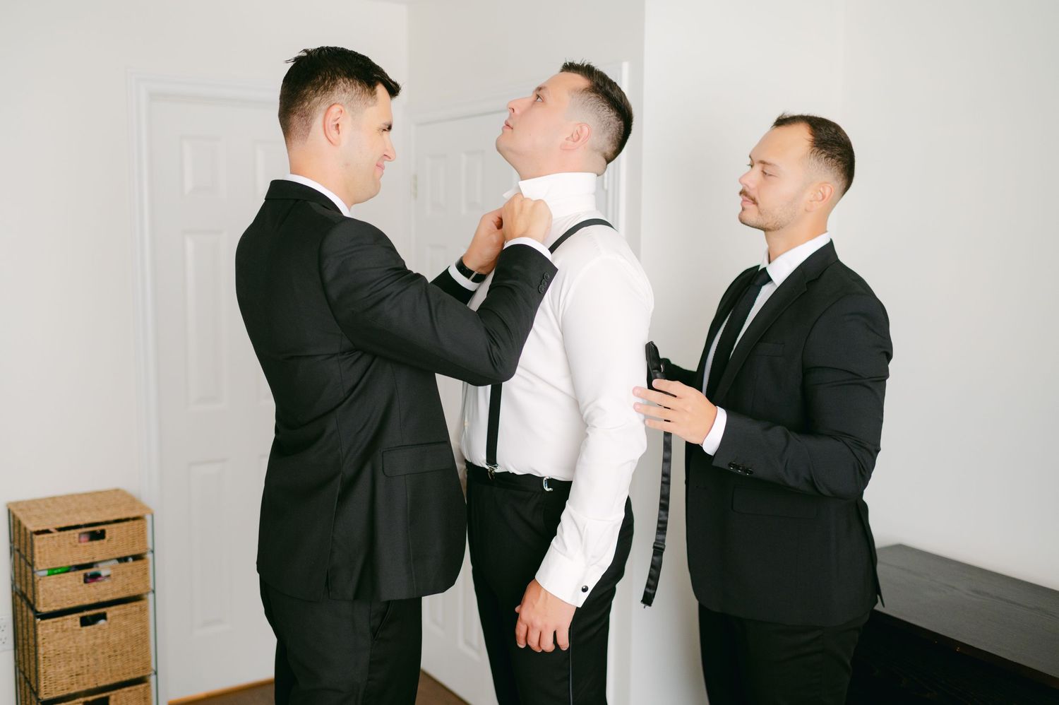 Three people in formal attire helping with wedding day preparations in a bright indoor setting.