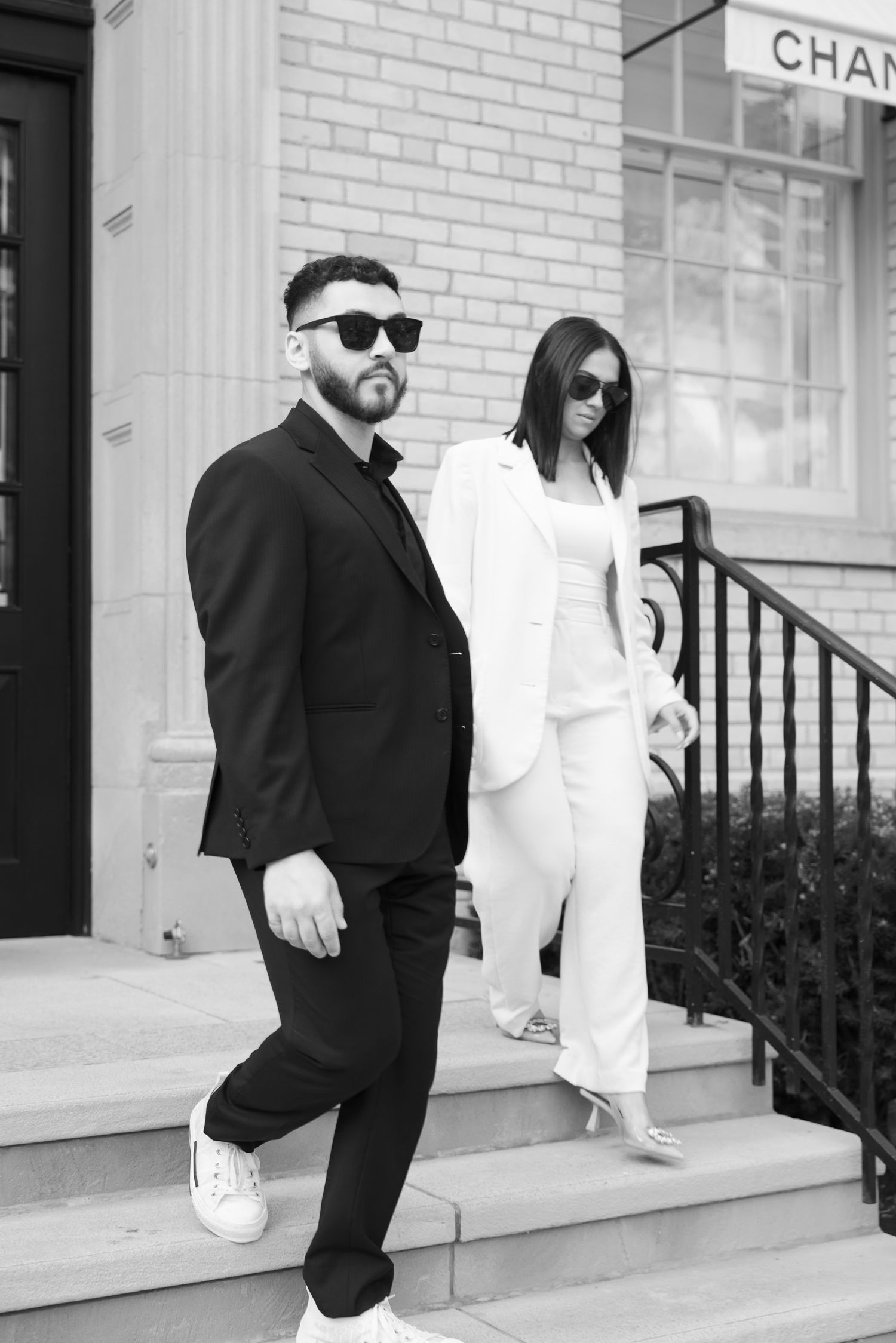 Stylish couple in contrasting black and white outfits pose on steps outside a Chanel storefront.