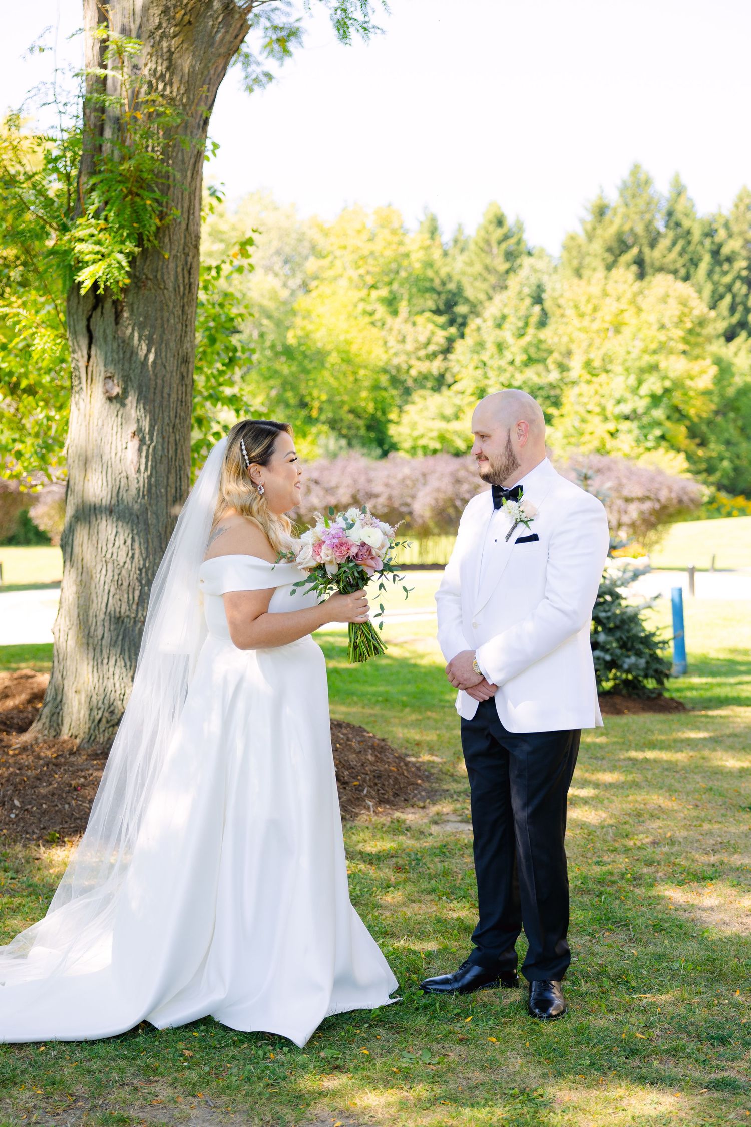 Elegant outdoor wedding scene with couple in white attire standing beneath a large tree.