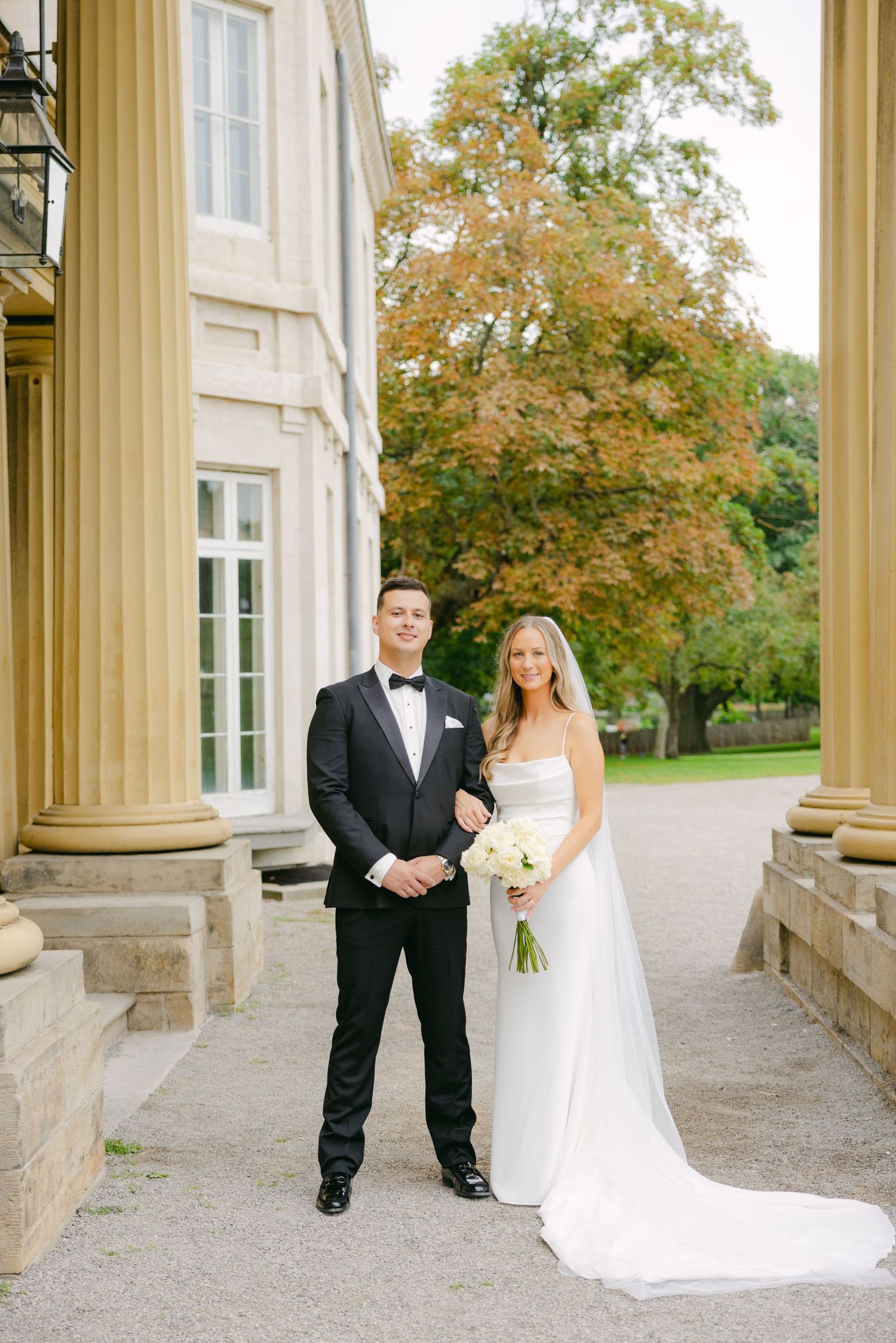 Full length wedding portrait with couple standing between classical columns in autumn.