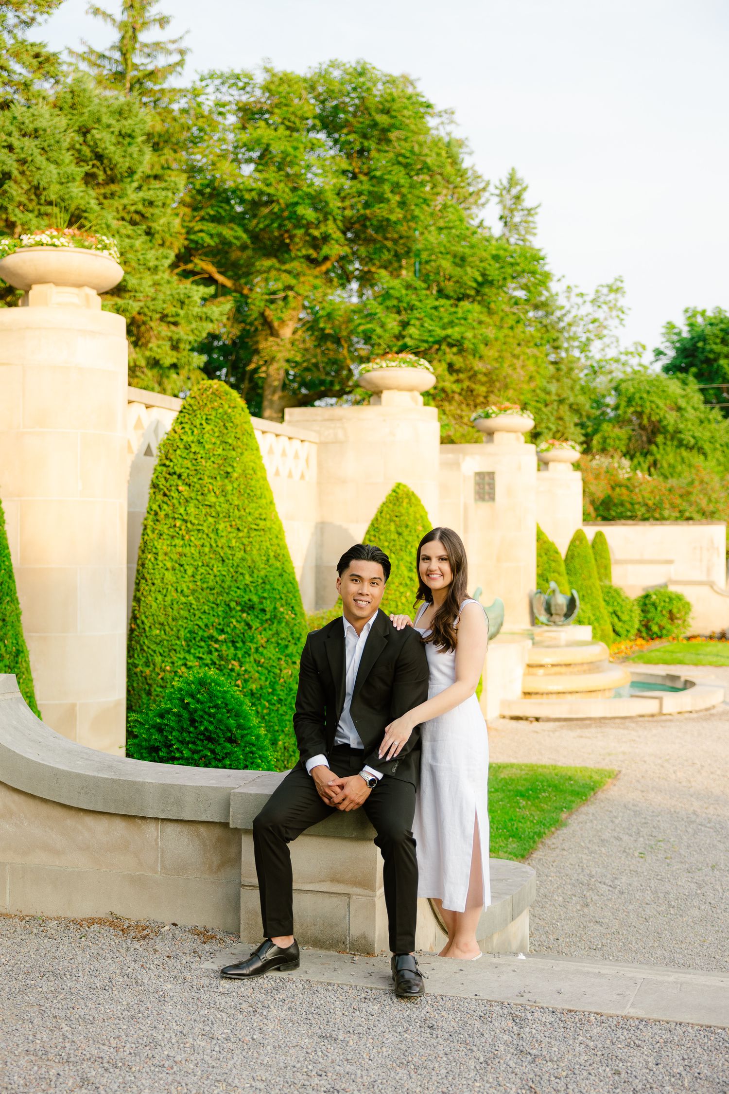 A couple posing together on a stone wall in a manicured garden with topiary trees.