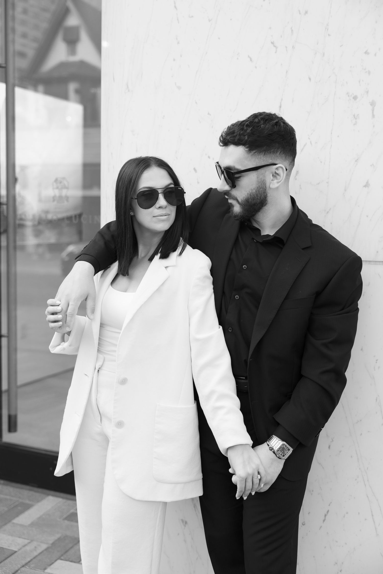 Black and white portrait of couple in formal wear sharing intimate moment against building wall.