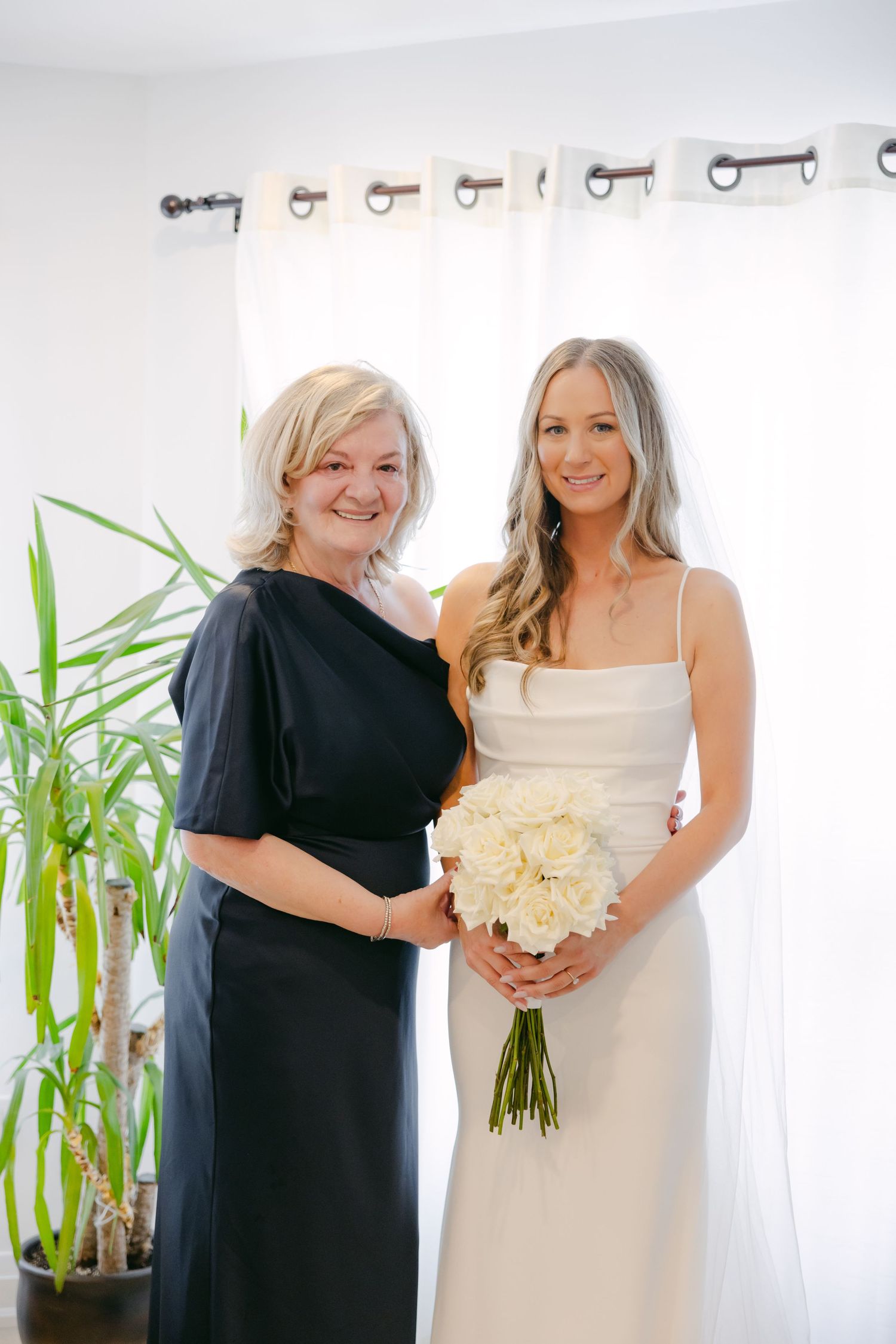 Bride in white dress holds bouquet of white roses while posing with woman in black dress.