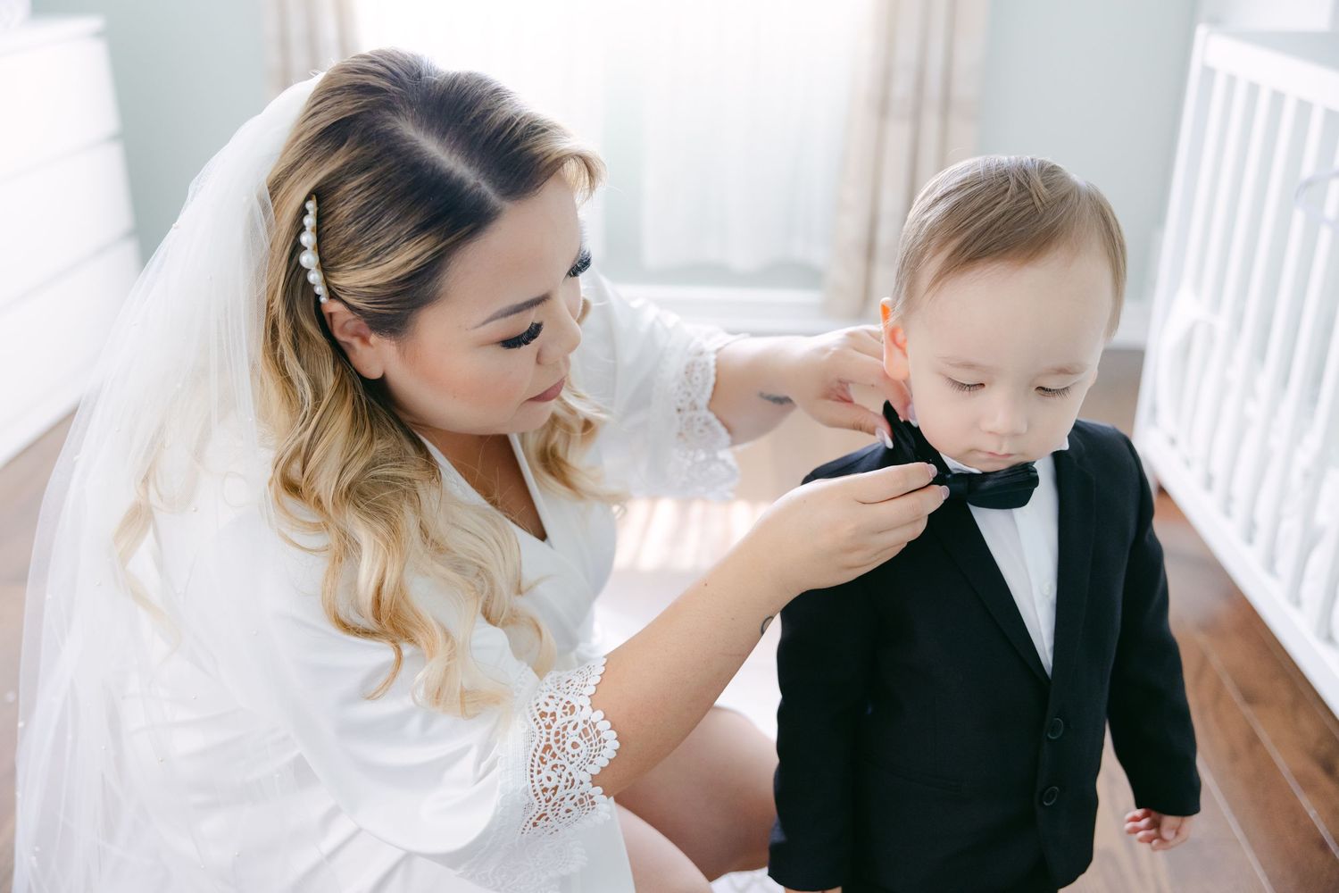 Mother in bridal attire adjusting toddler's bowtie on wedding day.