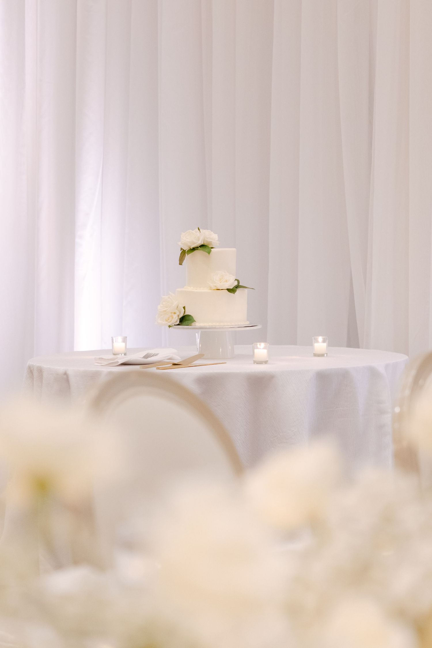 Three-tiered white wedding cake with floral accents displayed on round table with white linens and candlelight.