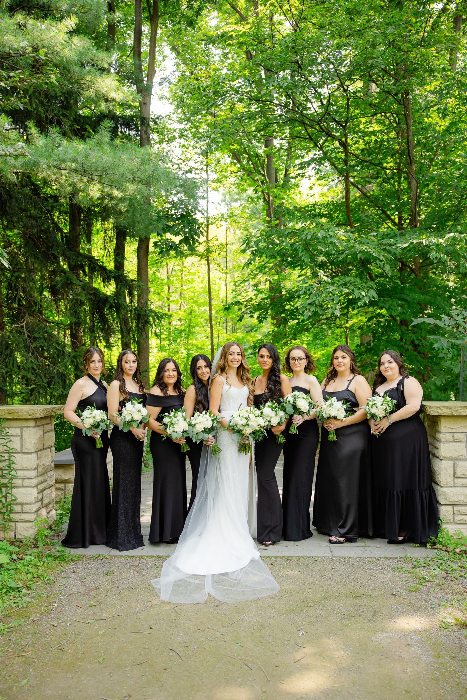 A formal wedding party photo with bridesmaids in black gowns holding white bouquets against forest backdrop.