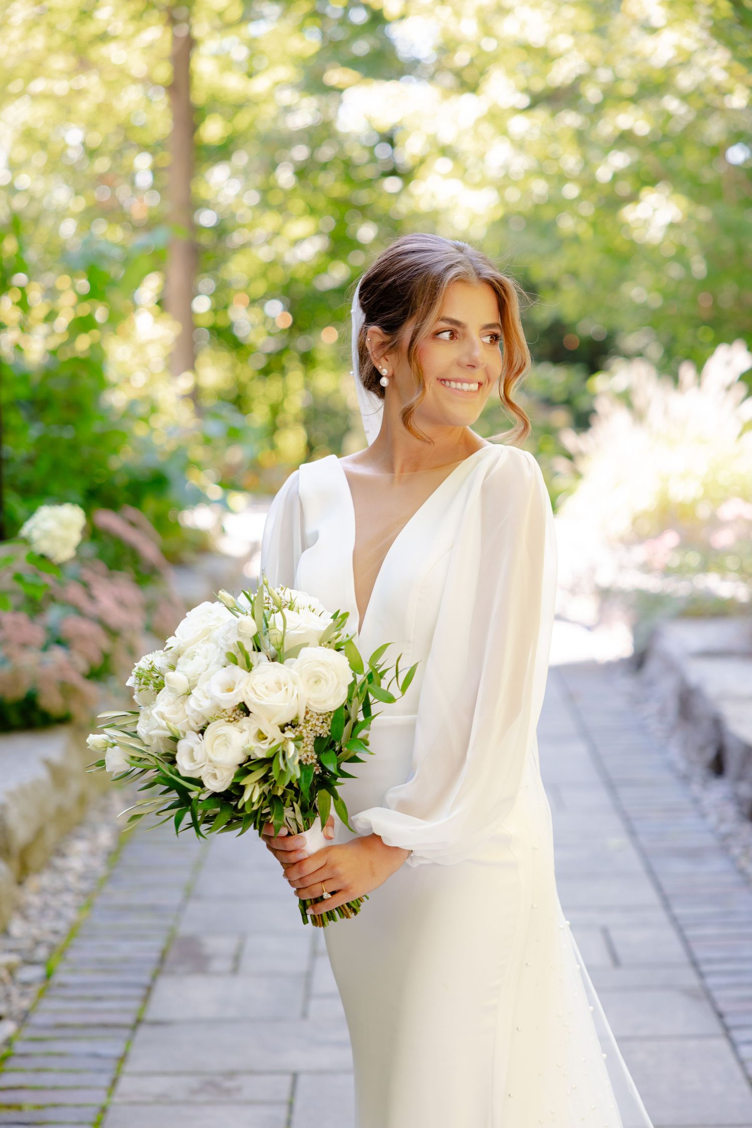 Bride in long-sleeved white gown holding elegant white floral bouquet in garden setting.