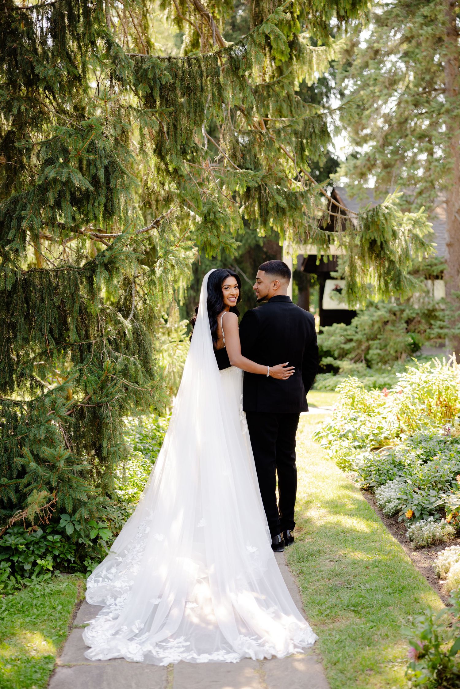 Wedding couple walks along a garden path under towering evergreen trees in romantic lighting.