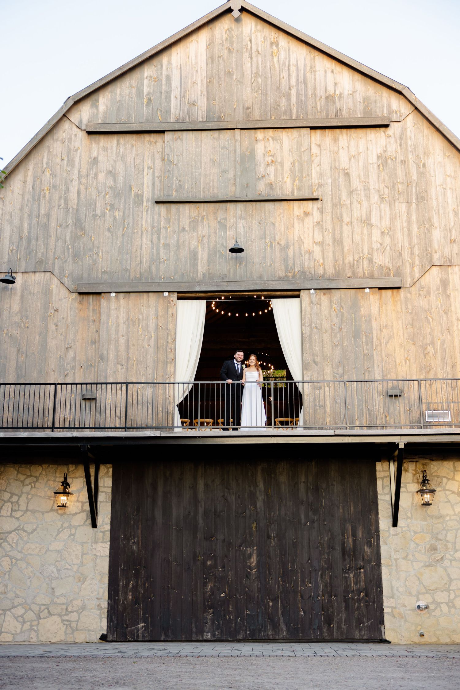 Couple standing on a rustic barn balcony framed by large wooden doors and string lights.