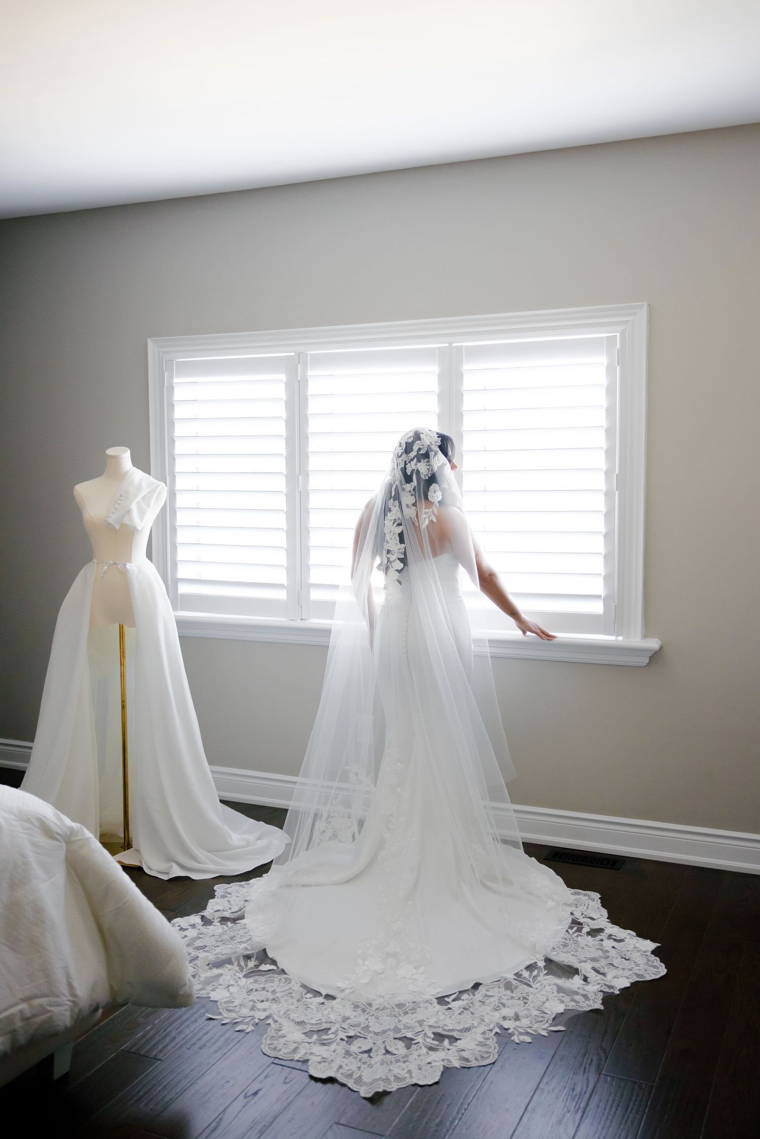 Elegant bridal gown and veil silhouetted against bright window with dress form in background.
