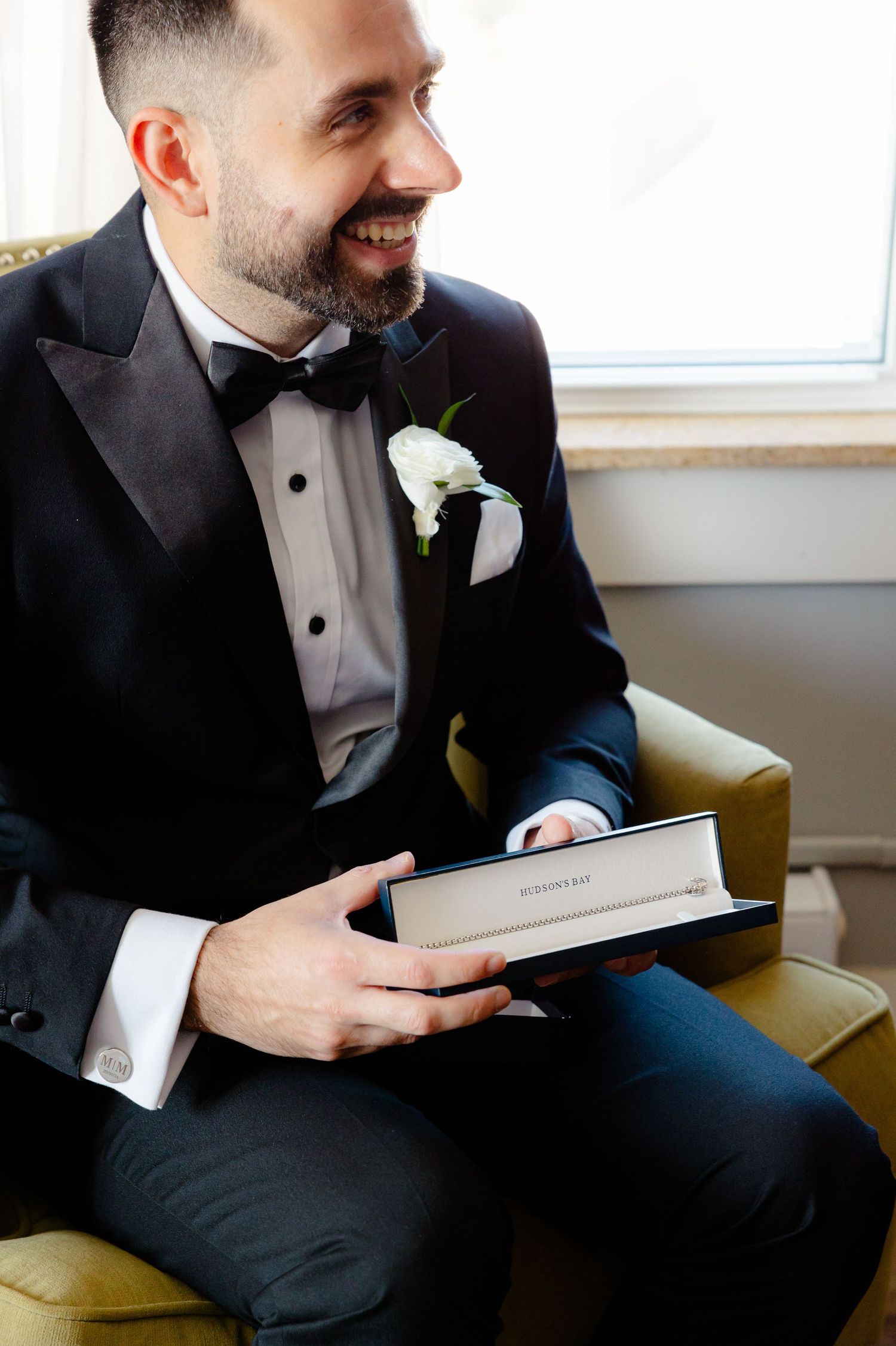 A person in formal attire smiles while holding a book in a vintage armchair.