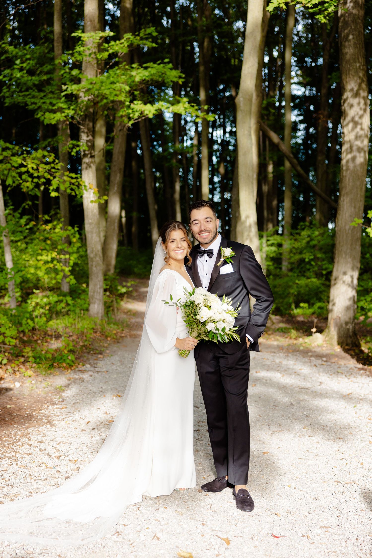 Elegant wedding couple poses on wooded path with bride holding white floral bouquet.