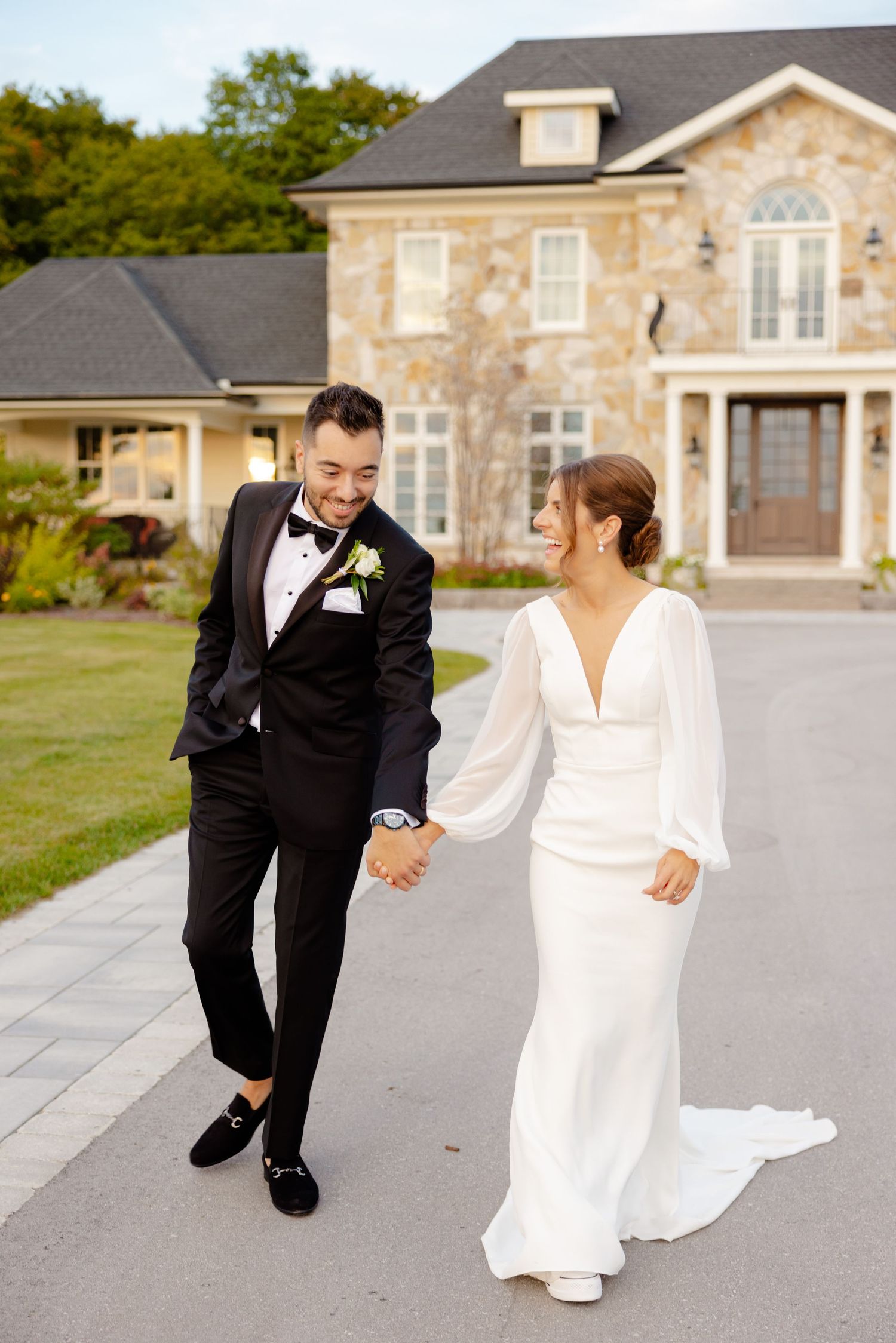 A couple in wedding attire strolling together on a path in front of an elegant stone mansion.