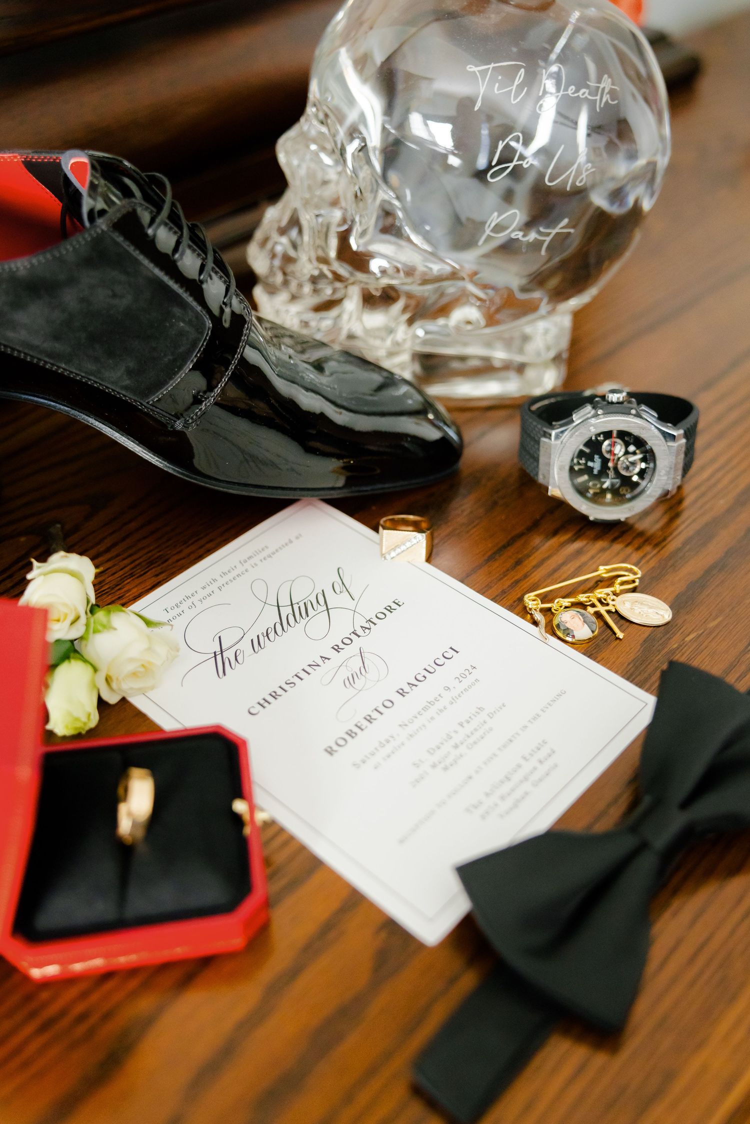 Wedding details flatlay with crystal skull, watch, shoes, invitation and bow tie on wooden surface.
